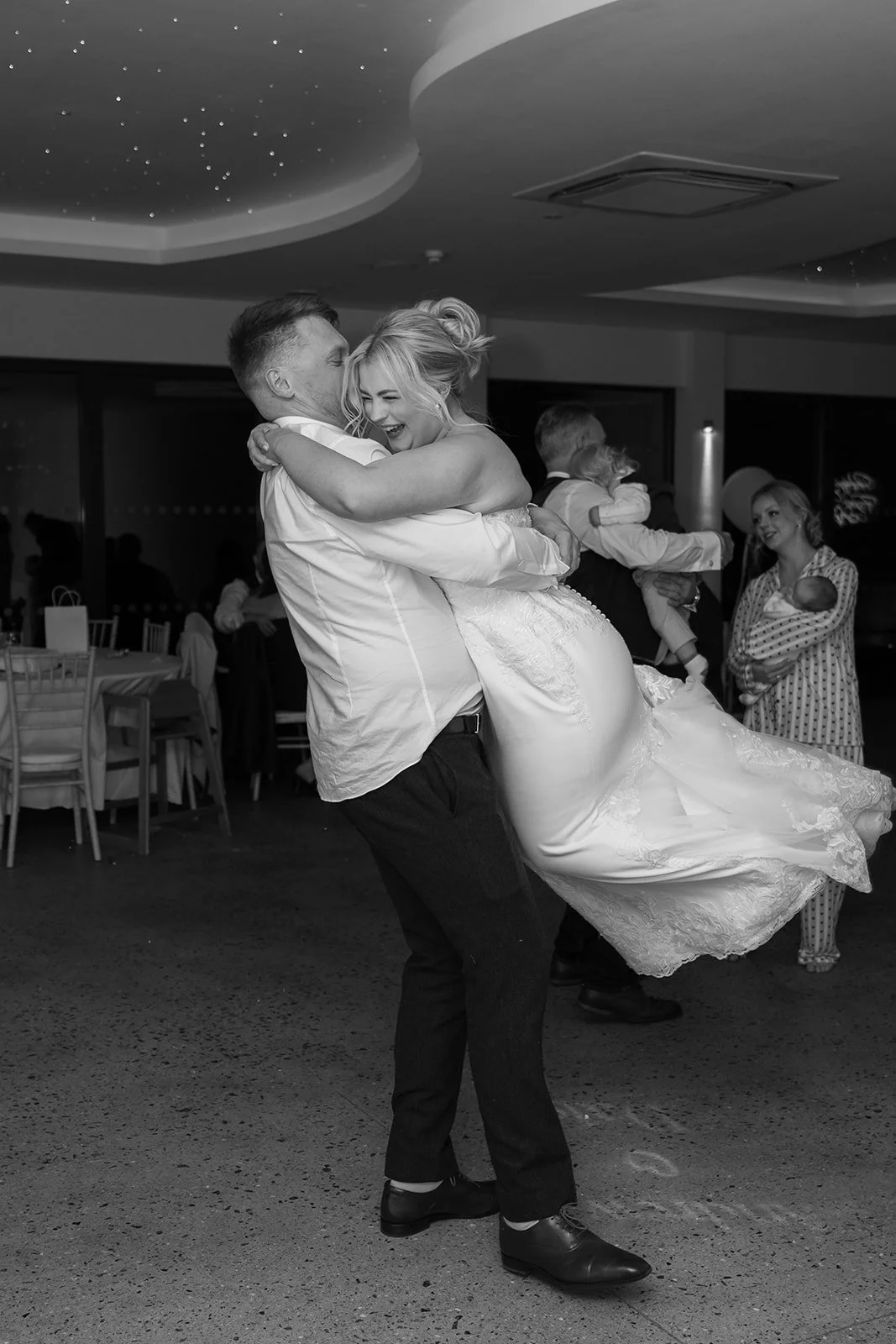 Andrew lifting Daisy in a joyful embrace on the dance floor at Sandy Cove Hotel in Devon, capturing candid evening reception celebrations with guests and twinkling ceiling lights at this modern coastal wedding venue.