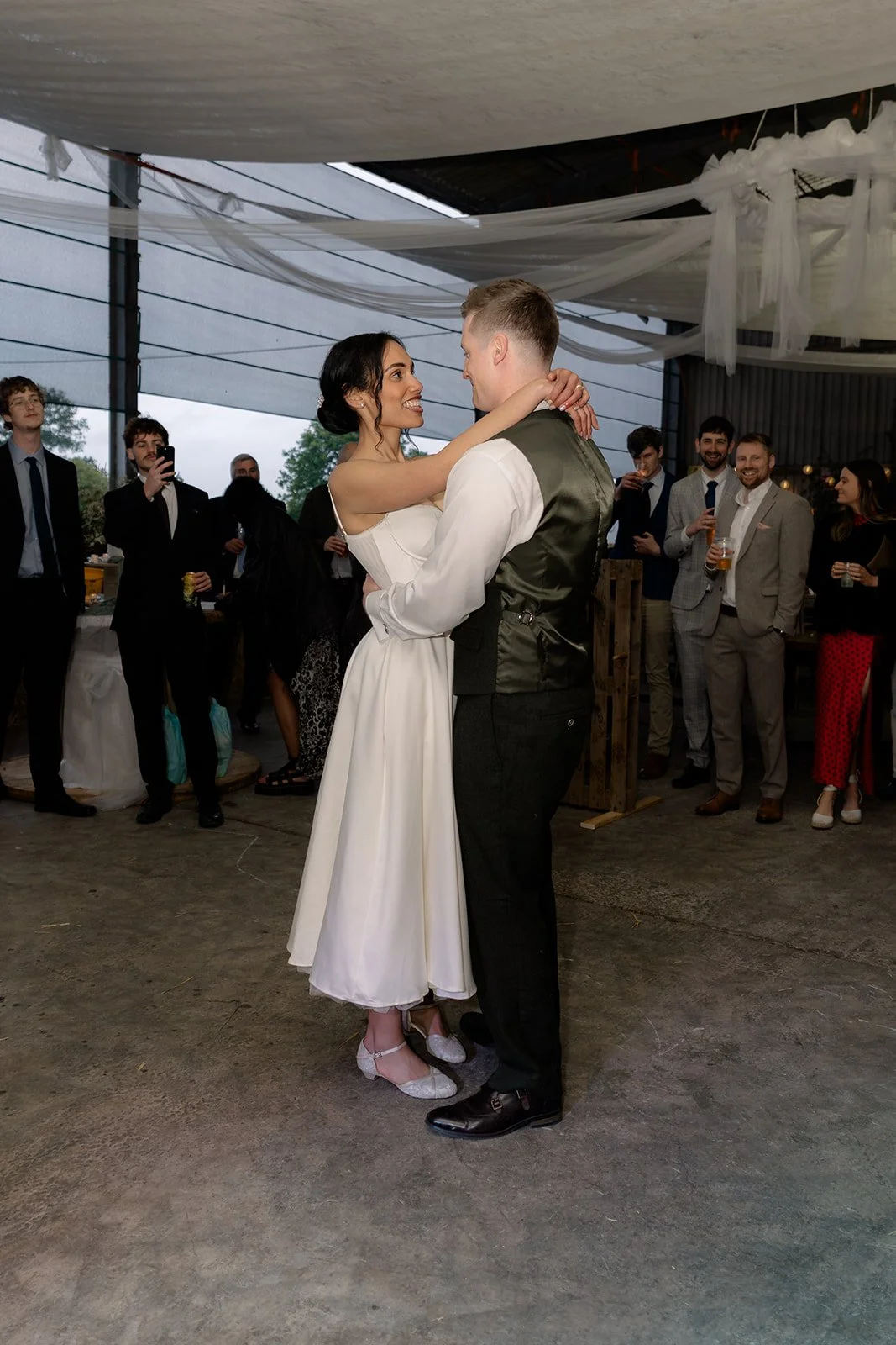 Jaye & Josh share their first dance at Farrington's Farm in Bristol, smiling together beneath soft draped fabric as guests watch nearby, with rustic concrete flooring, relaxed barn reception details and easy evening celebration atmosphere now.