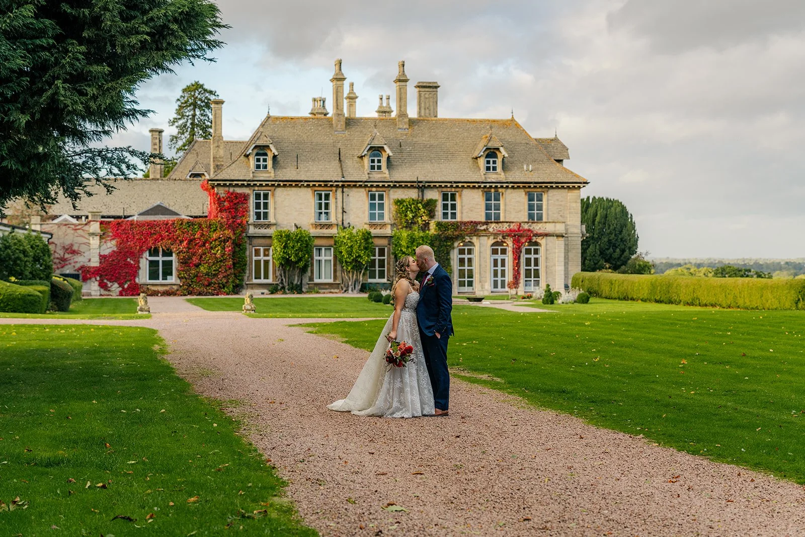 Lucy & Jay kissing on the lawn at Eastwood Park in Gloucestershire, with the manor house behind them, photographed by Just James Weddings for relaxed wedding portraits.