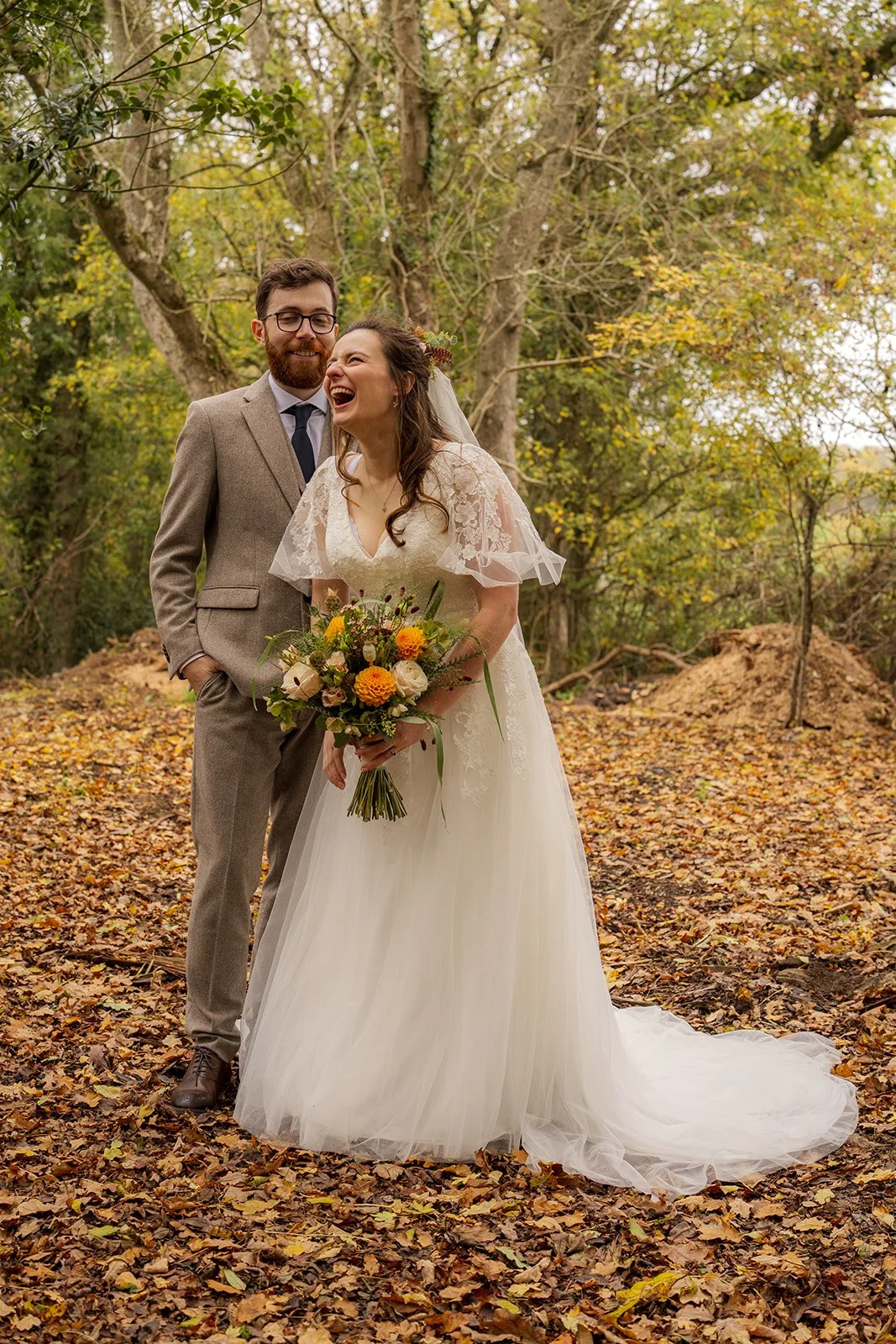 Maisie & Andrew laughing together in autumn woodland at Stockbridge Farm Barn in Dorset, surrounded by fallen leaves and warm seasonal colours, photographed by Just James Weddings.