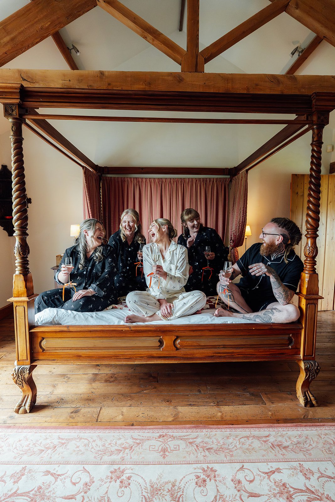 Candid morning photo of Hannah & Ben’s wedding party laughing on a four-poster bed at Grays Barn in Devon, UK, holding drinks in matching pyjamas during relaxed bridal preparations in the room, photographed by Just James Weddings.