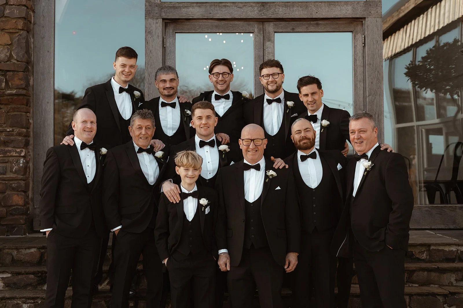 Jay poses with his groomsmen outside Upton Barn & Walled Garden in Devon, UK. The group wears classic black tuxedos, sharing relaxed smiles and connection during Jessie & Jay’s wedding day group portraits.