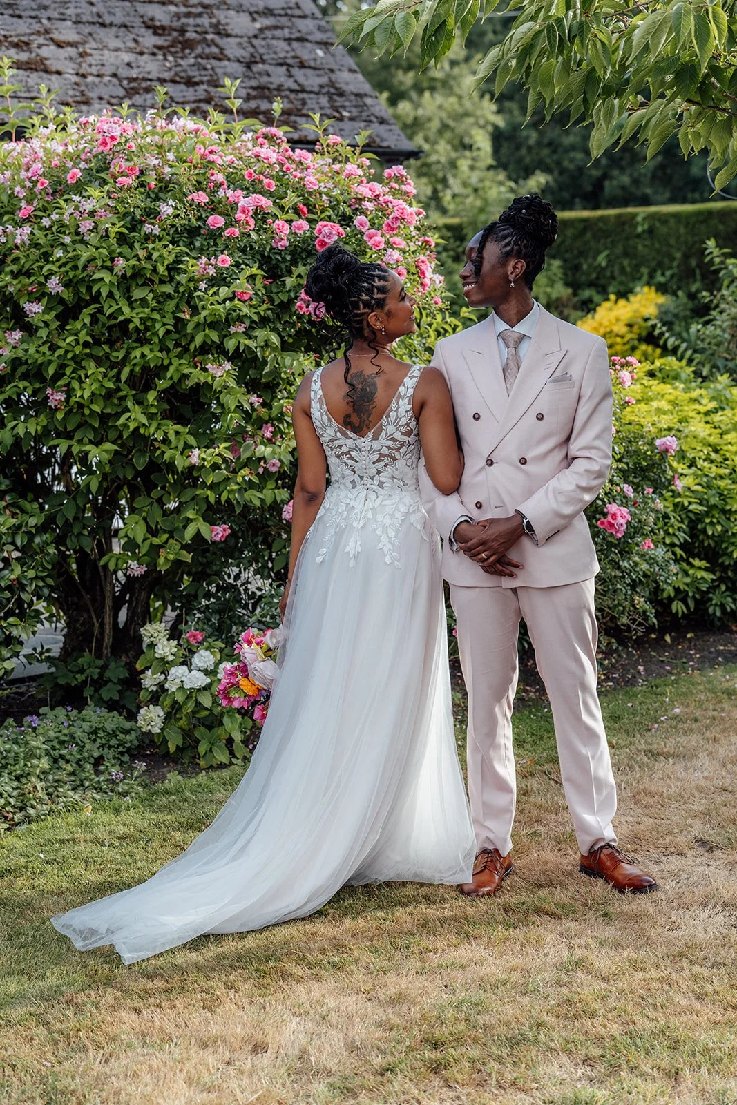 Kasey and Larisha sharing a relaxed moment beside blooming pink flowers during their wedding portraits in the gardens at Oaks Farm Weddings in Surrey.