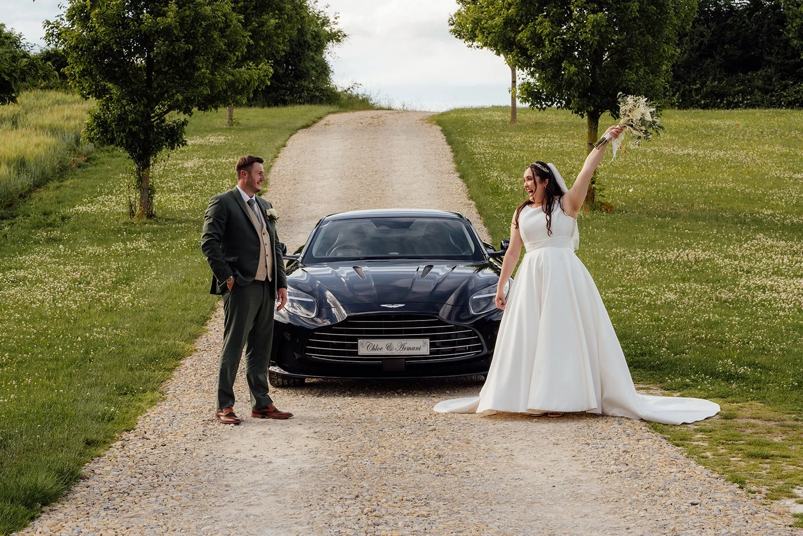 Chloe & Armani posing with a black sports car on the country drive at The Barn at Upcote in the Cotswolds, photographed by Just James Weddings for fun wedding portraits.
