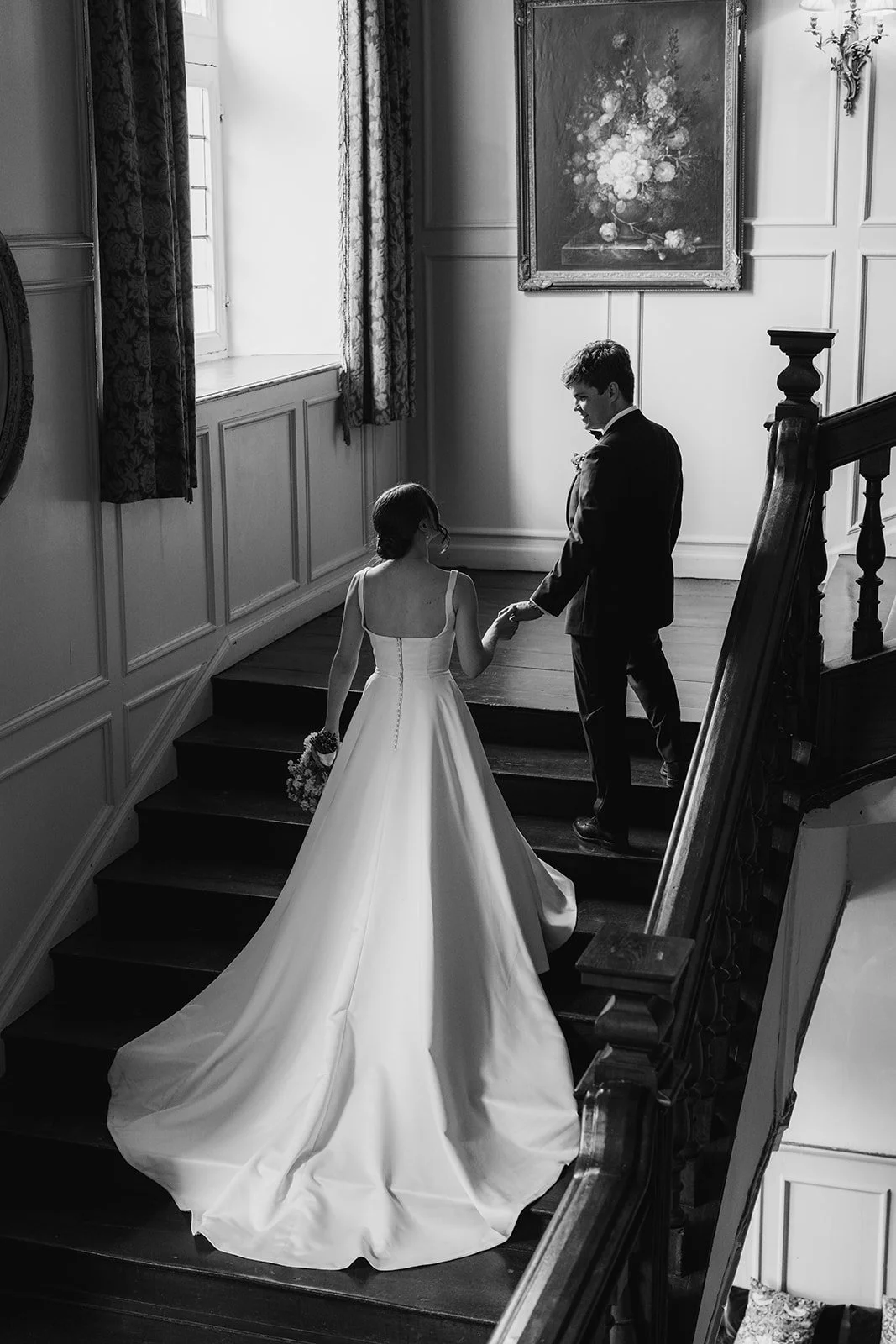 Black and white photo of Rachel & Tom walking hand in hand up the grand wooden staircase inside Brympton House in Somerset, photographed by Just James Weddings.