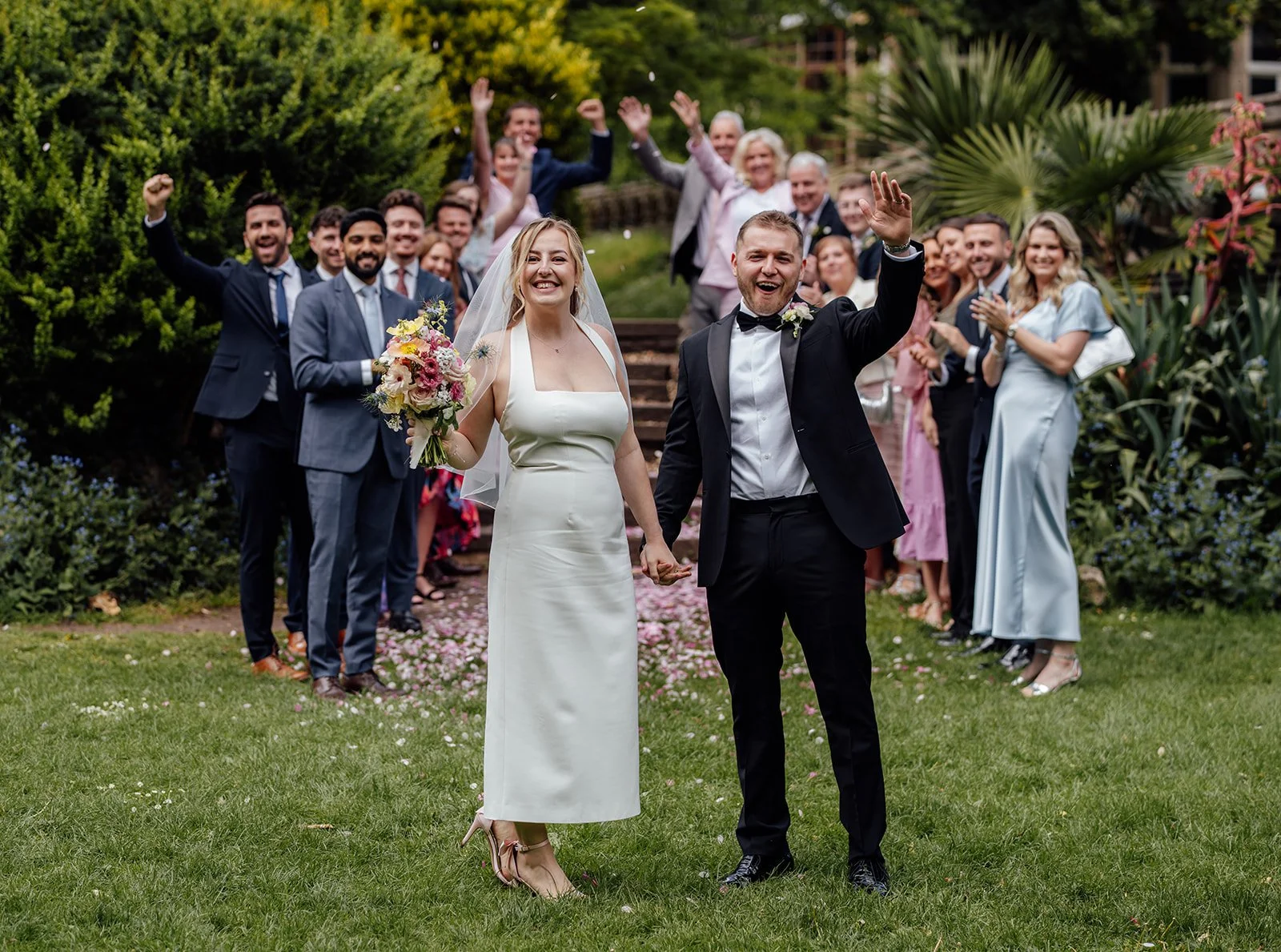 Bryony & Max stand smiling together on the lawn at Insole Court in Cardiff, holding hands as their guests cheer behind them, with confetti on the path, garden greenery all around and Bryony holding her colourful wedding bouquet nearby together.