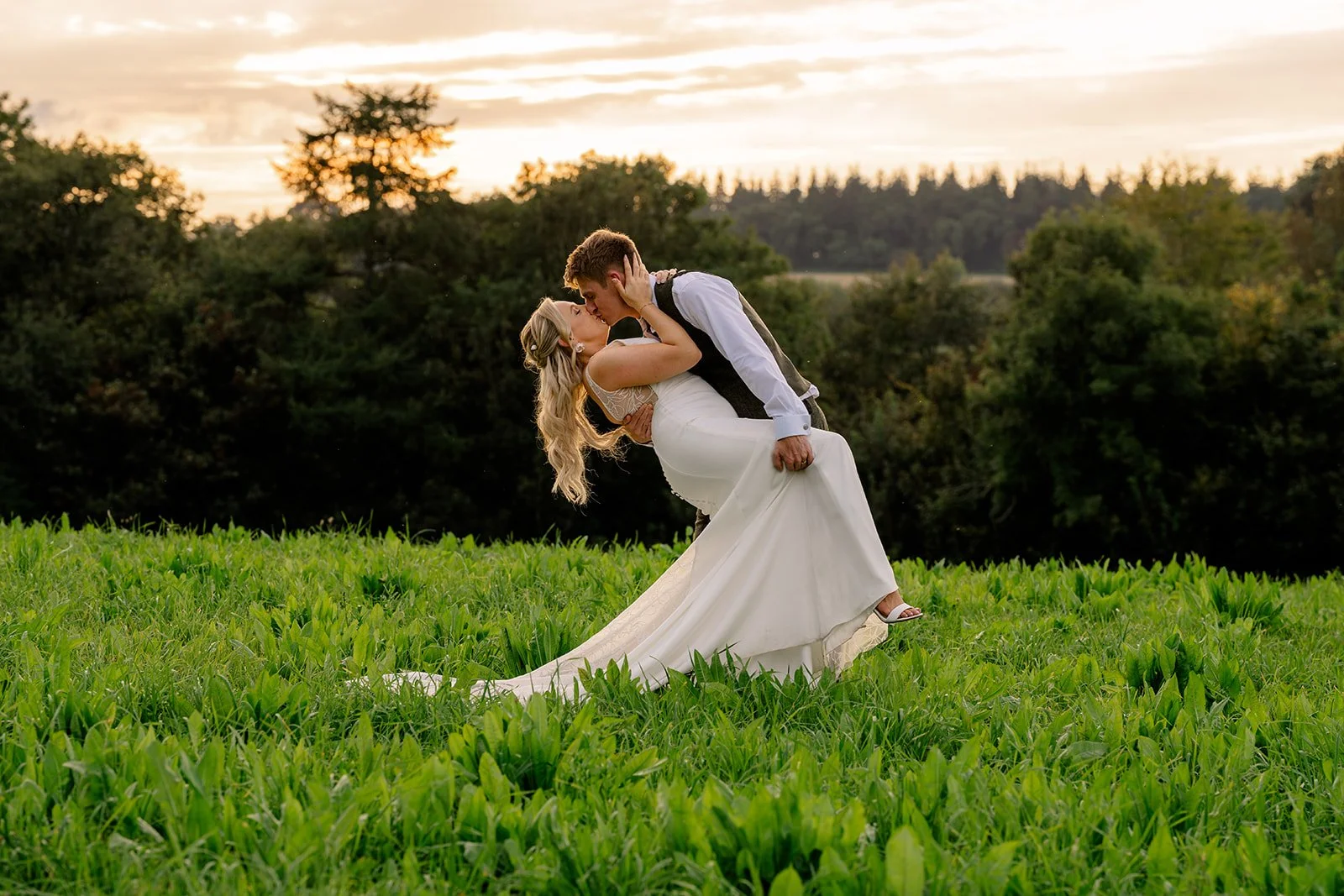Abi & Josh sharing a sunset kiss in a field at The Cider Barn in Somerset, with the groom dipping the bride against soft evening light, photographed by Just James Weddings for romantic countryside wedding portraits.