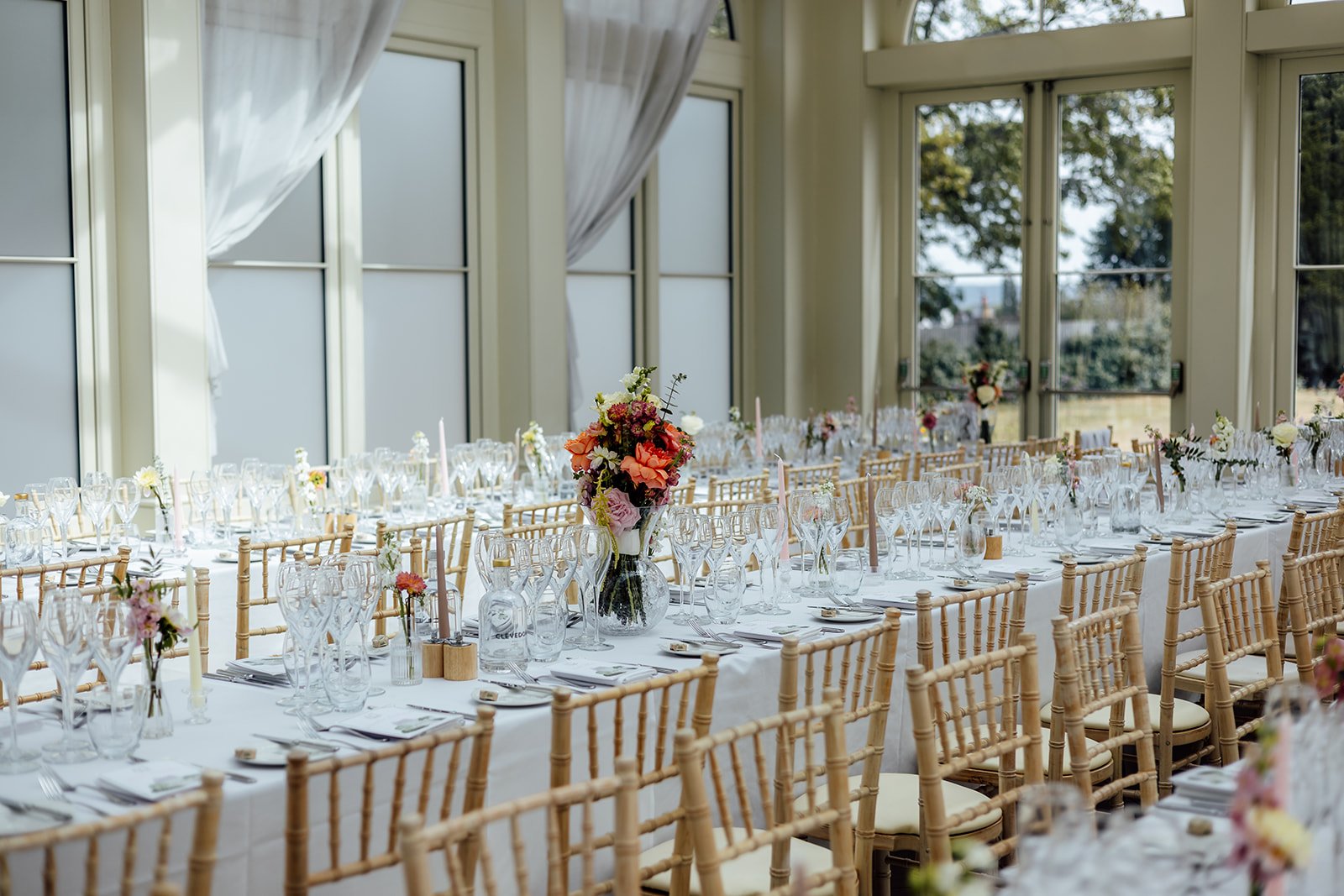Wedding breakfast tables at Clevedon Hall in Somerset, with gold chairs, floral centrepieces and glassware beside tall windows, photographed by Just James Weddings.