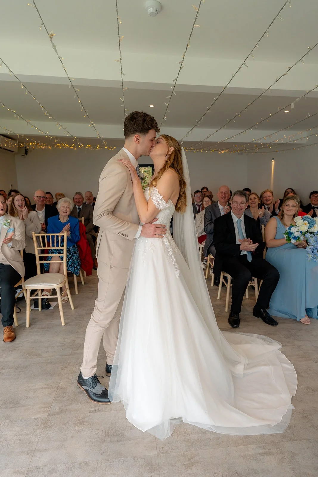 Hannah & Cameron share their first kiss as newlyweds during their wedding ceremony at Sandy Cove Hotel in North Devon. Guests smile and applaud as the couple celebrate their first married moment inside the bright coastal ceremony room.