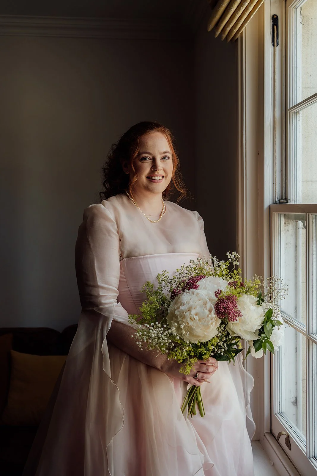 Charlotte & Jack wedding portrait at Bishop’s Palace in Somerset, UK, with the bride in a soft pink dress holding a white and pink bouquet by a tall window, lit by natural light during calm morning preparations before the ceremony in Wells.
