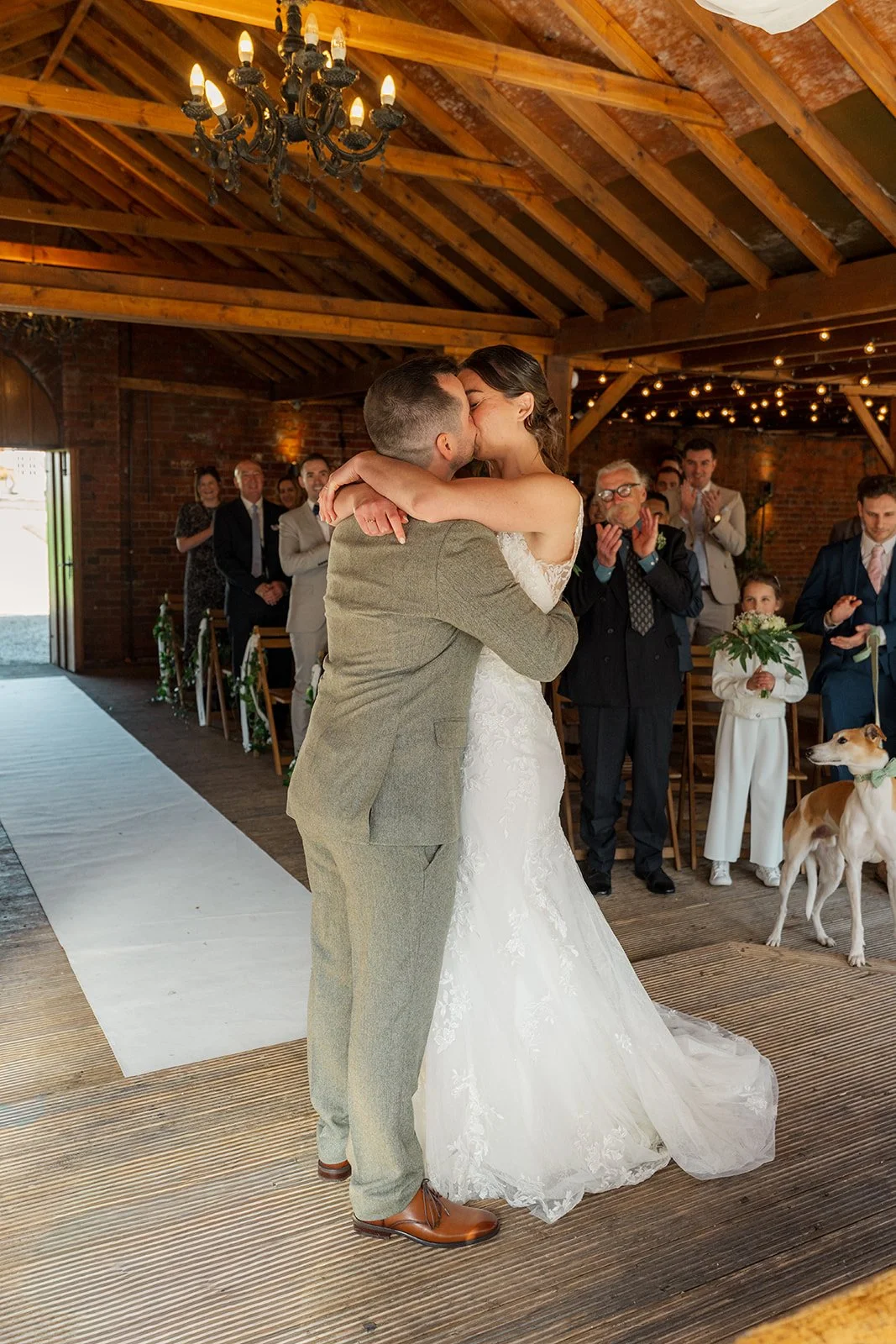 Chris and Anna share their first kiss during the ceremony at Elmhay Park in Somerset, UK, as guests stand and applaud inside the rustic barn and their dog watches nearby. A heartfelt just-married moment from Anna & Chris’s wedding day together.