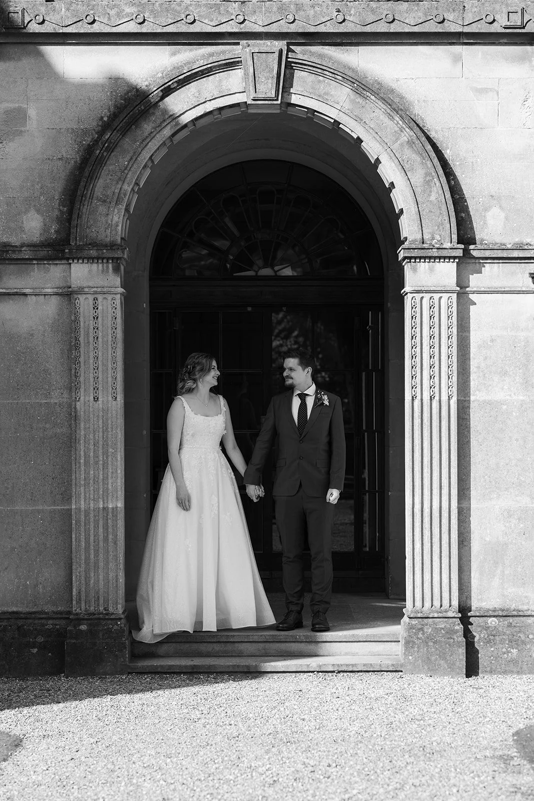 Black and white wedding portrait of Jessie and Thomas holding hands beneath an arched doorway at Coombe Lodge in Somerset, UK, sharing a quiet moment together.