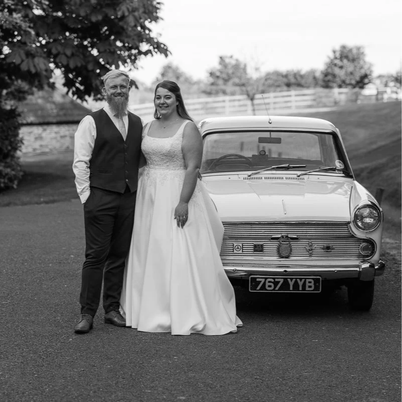 A bride and groom standing next to a vintage car outdoors. The groom has a beard and is wearing a vest and tie, while the bride is in a wedding dress. Trees and a fence are in the background.