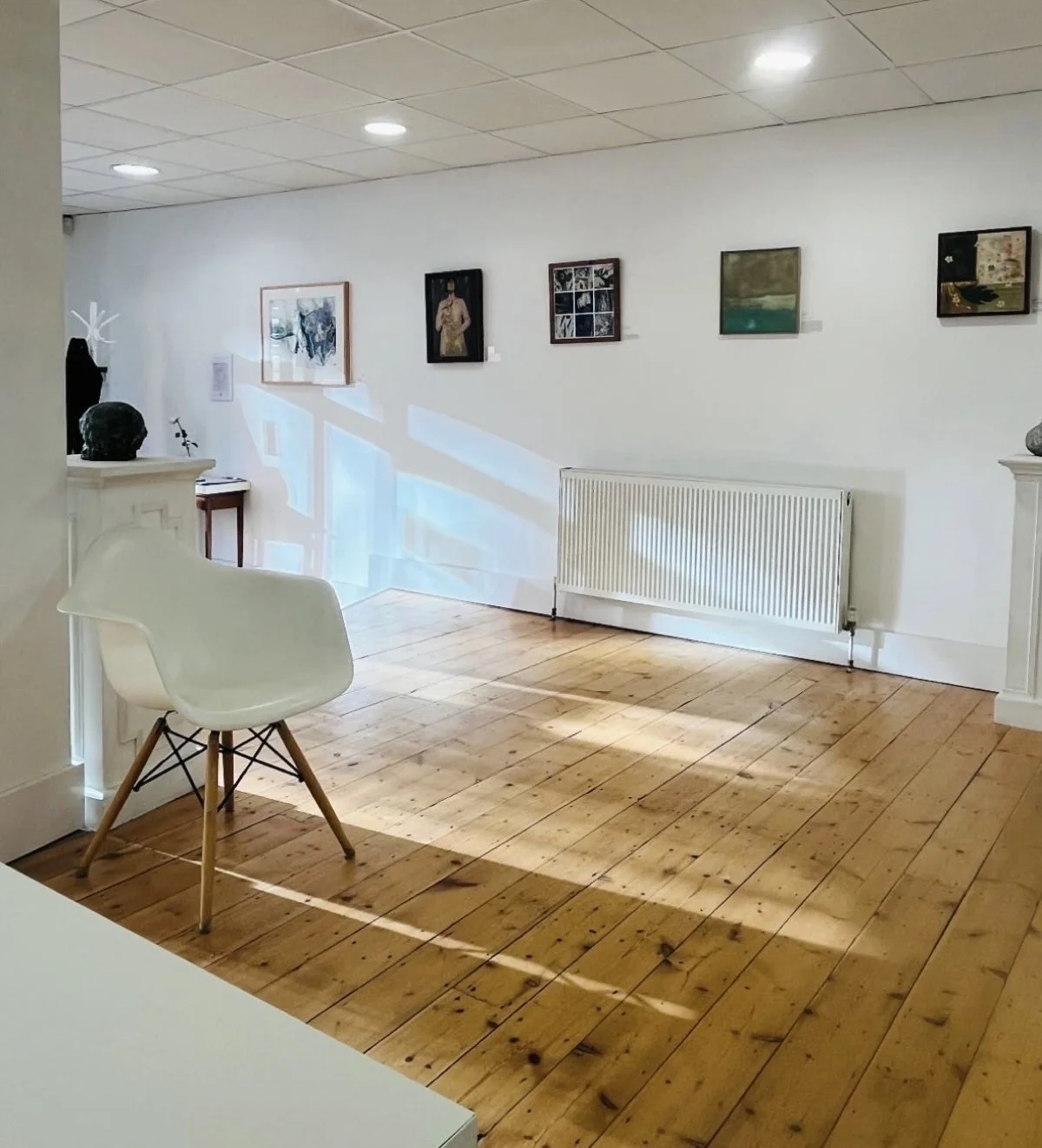 art exhibition called 'For Women By Women 6' with five paintings hanging on white wall and sunlight and shadows falling on wooden floor