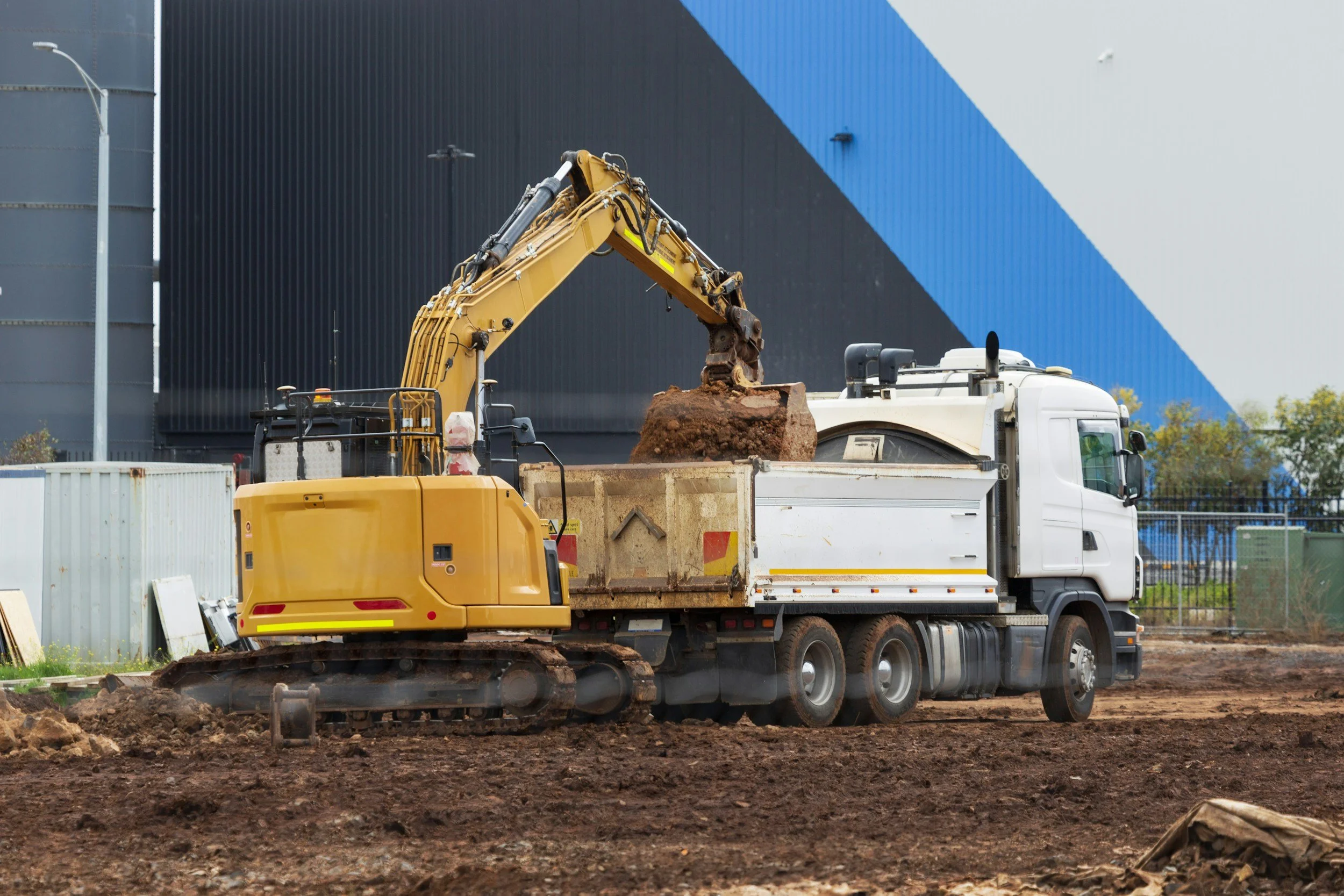 A construction site with a yellow excavator loading dirt into a white dump truck, with a large building in the background painted with black and blue geometric patterns.