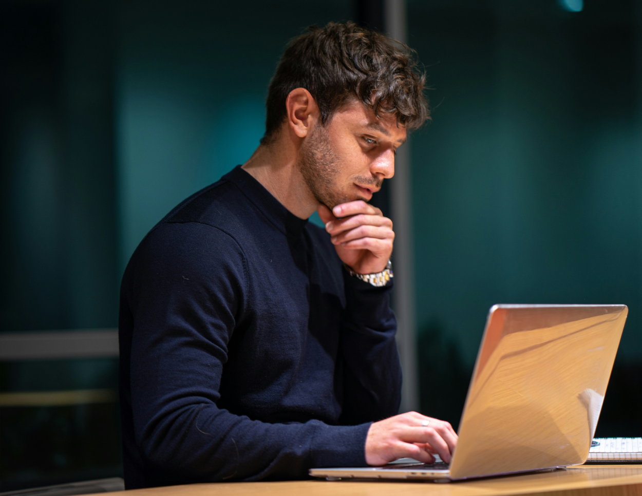 A man with brown hair sitting at a laptop, hand to chin with a pensive look on his face