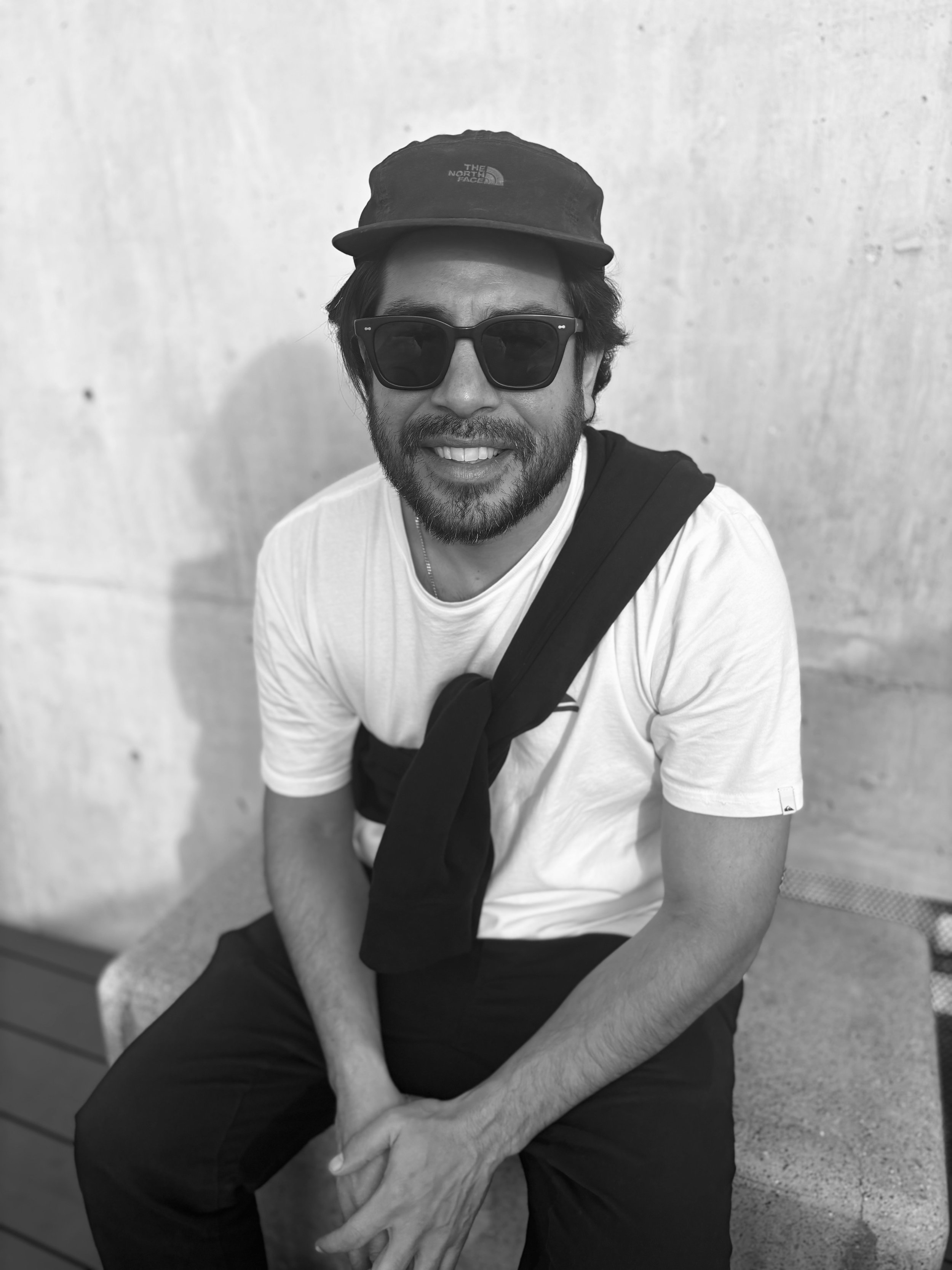 A smiling man with dark hair, a beard, and a mustache, wearing a black t-shirt and a silver chain, standing against a concrete wall.