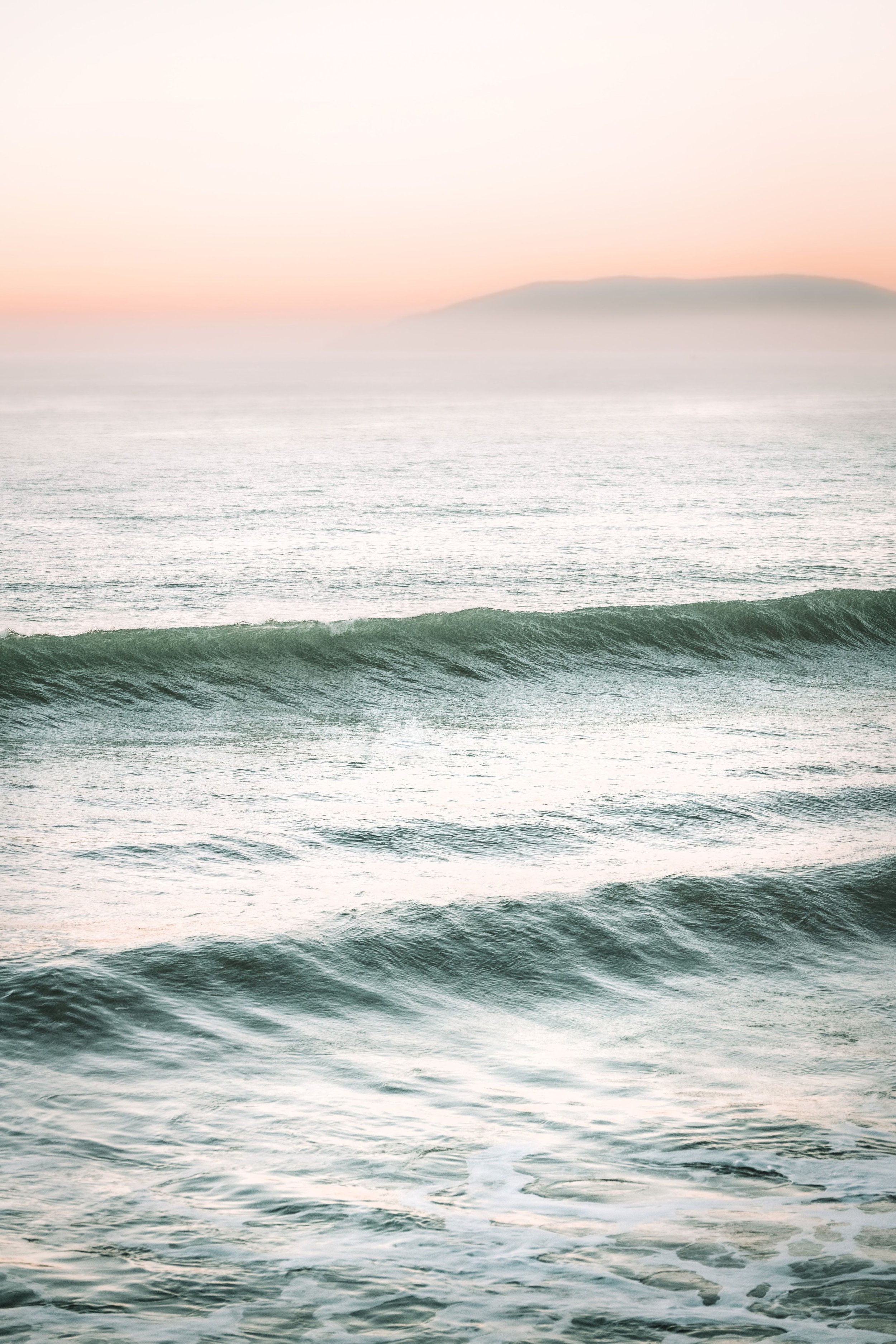 Calm ocean waves during sunrise or sunset with misty horizon and distant landmass.