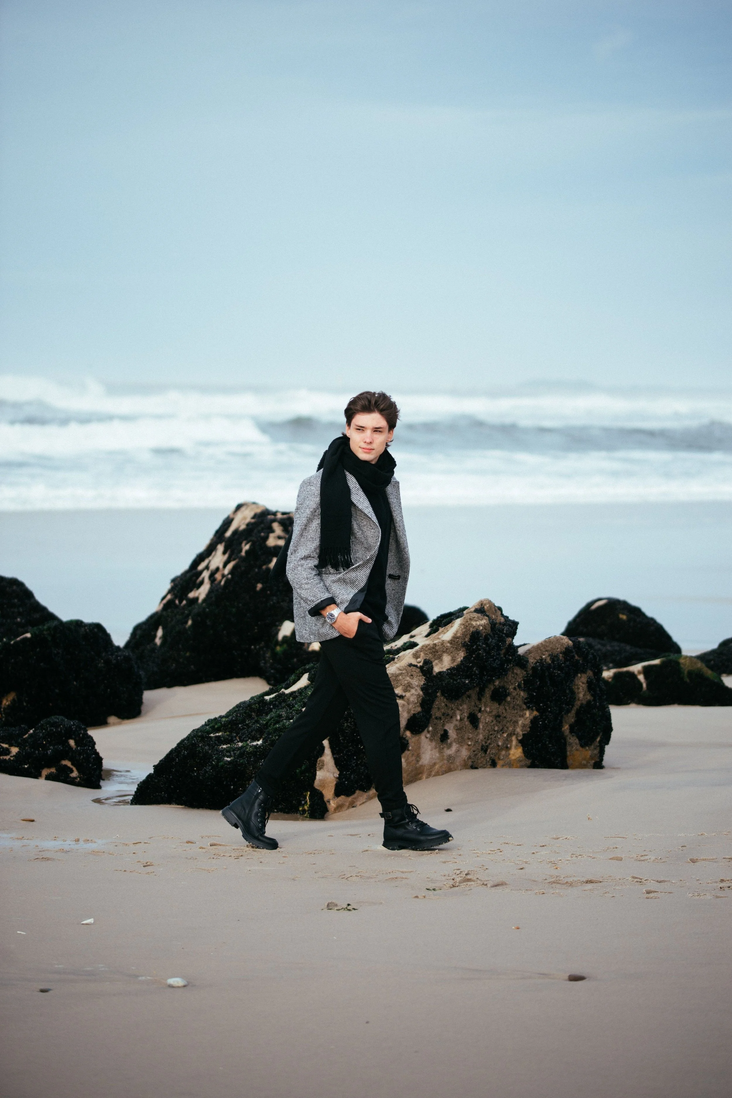 A young man walks on the beach wearing a gray coat, black scarf, black pants, and black boots, with rocks and ocean waves in the background.