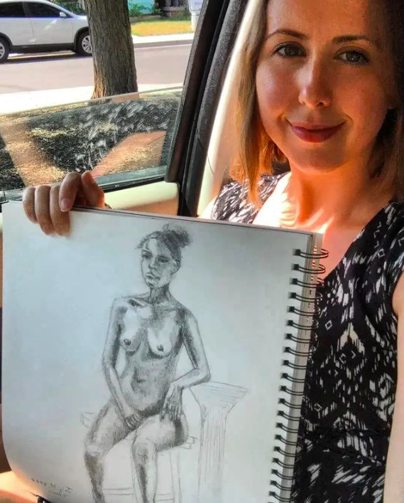 A woman sitting in a car holding a sketchbook with a pencil drawing of a nude figure.