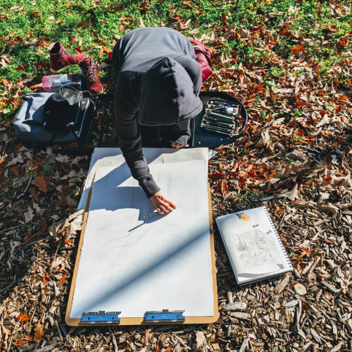 Person in a hoodie drawing outdoors on a large sketchpad, surrounded by art supplies and fallen leaves on the ground.
