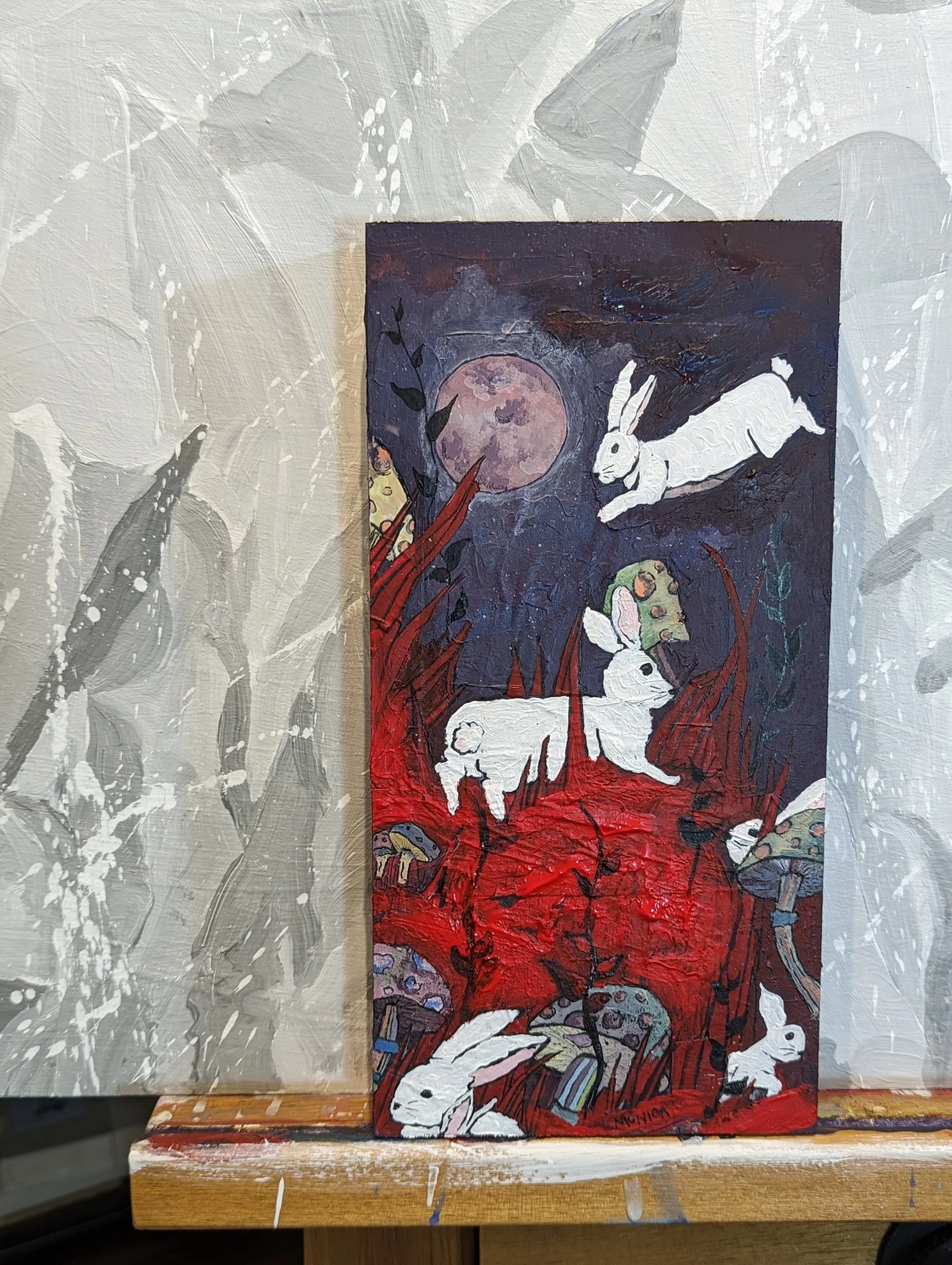 Painting depicting white rabbits jumping around red and purple foliage with a full moon in the background. The scene includes colorful mushrooms and dark atmospheric tones. The artwork is displayed on an easel against a gray, textured wall.