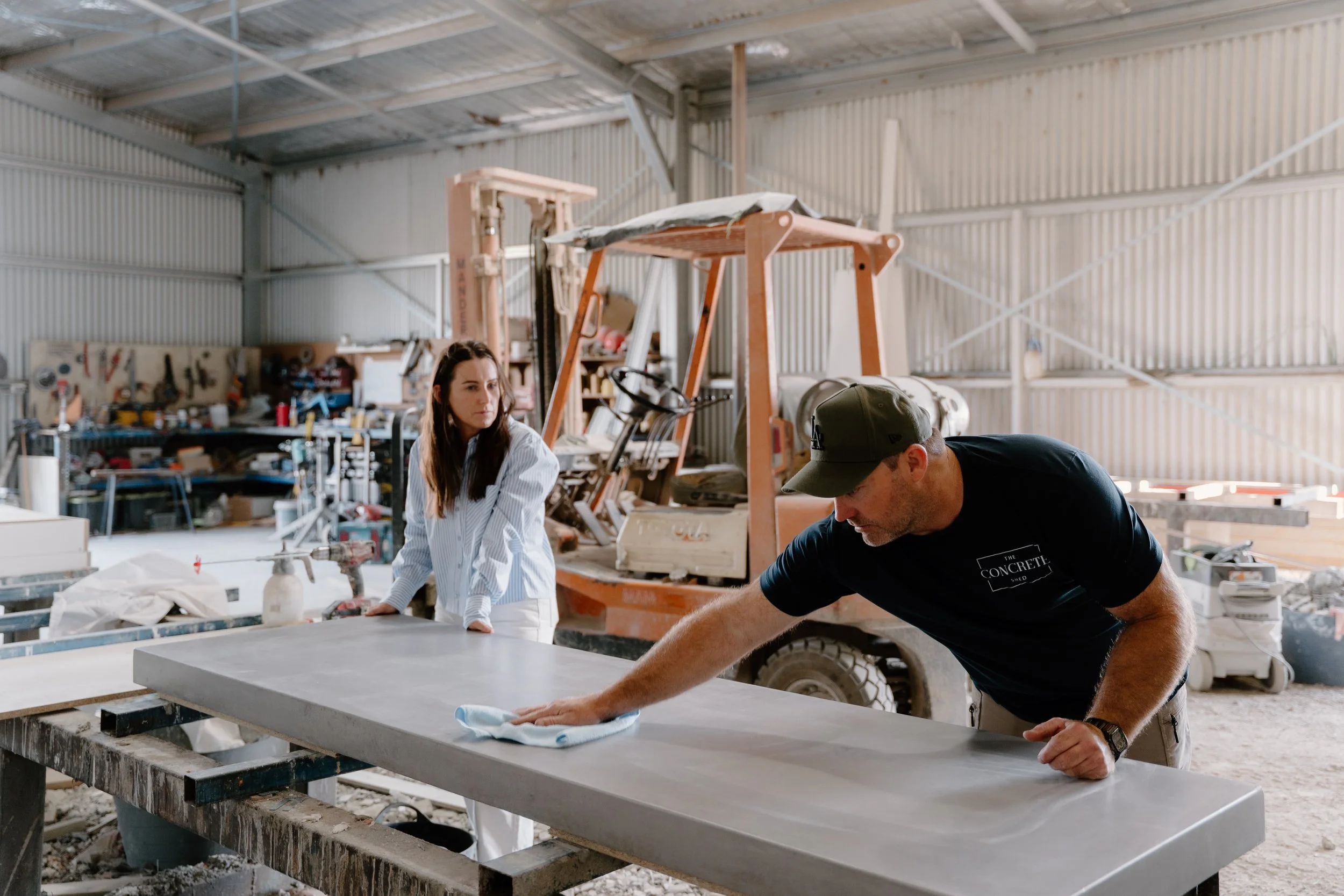 Two people working on a large concrete slab in a workshop. A man is cleaning the slab with a cloth, while a woman watches. The workshop has tools, equipment, and a forklift in the background.