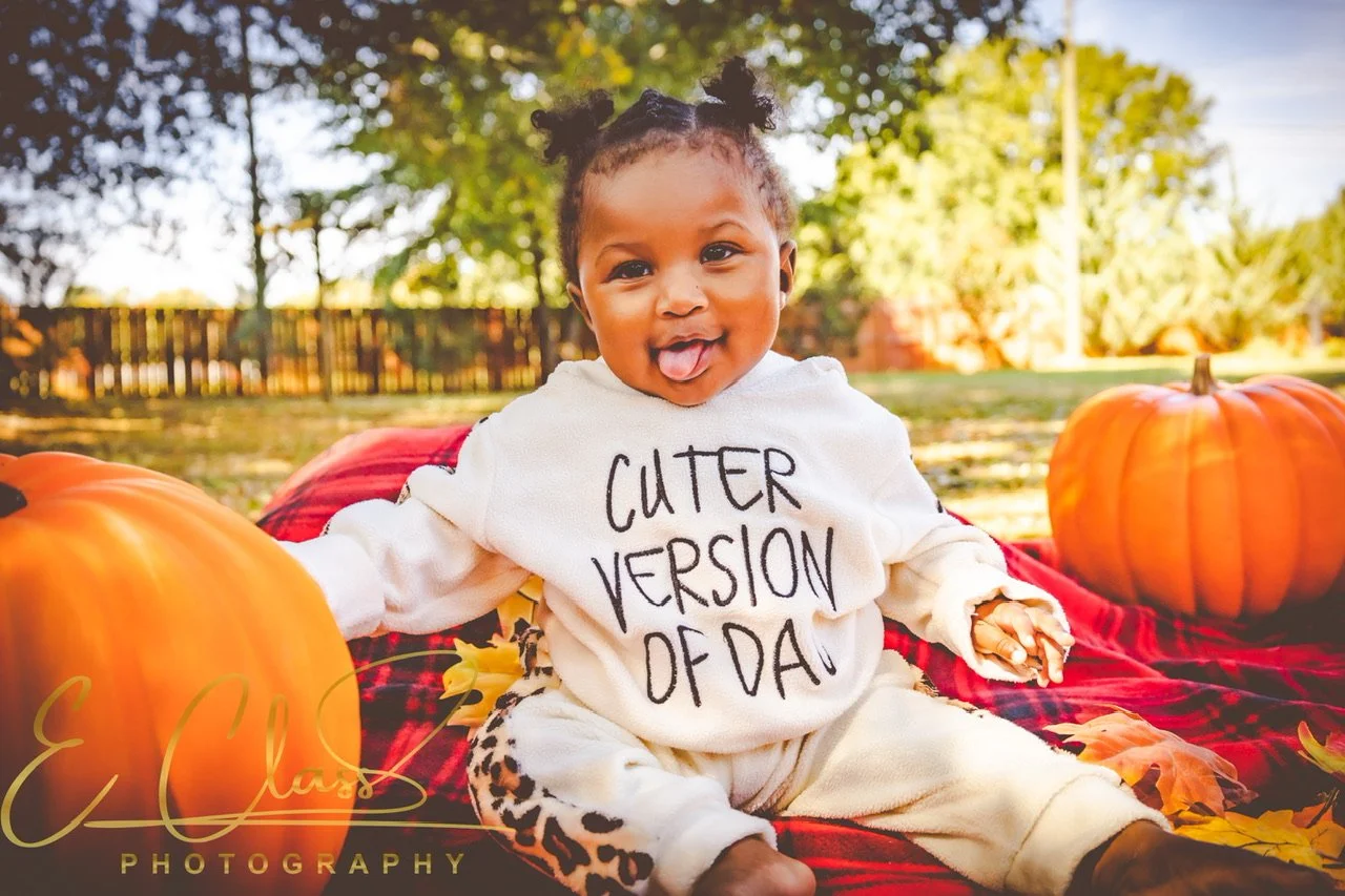 Smiling baby sitting outdoors with pumpkins, wearing a "Cuter Version of Dad" sweater, on a blanket surrounded by autumn leaves.