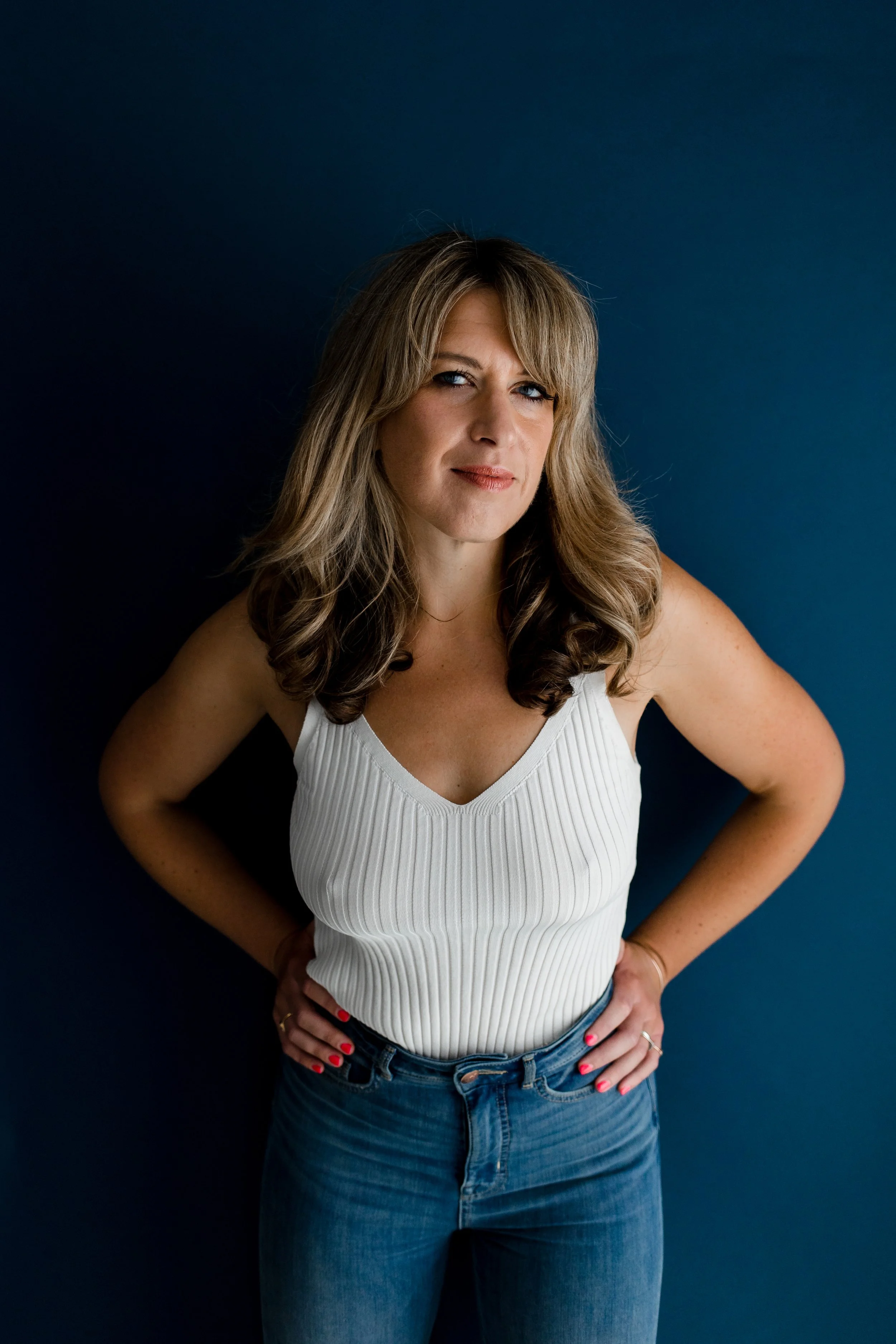 Daisy Buchanan is wearing blue jeans and a white vest and standing with her hands on her hips, smiling