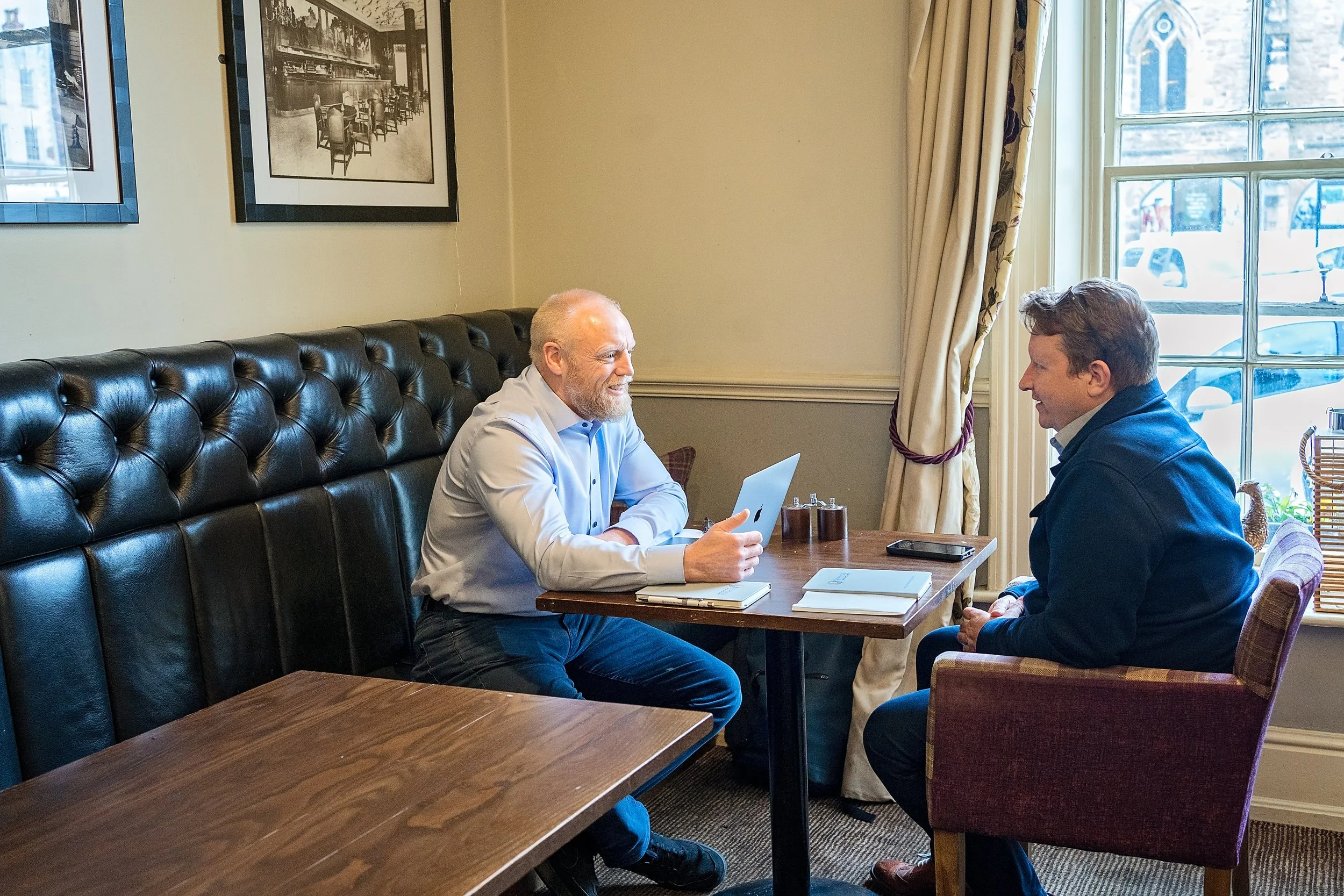 Two men are sitting at a table in a cafe, engaging in a conversation. One man has a laptop and is smiling, the other is listening. The cafe has framed pictures on the wall, large windows with curtains, and is well-lit with natural light.