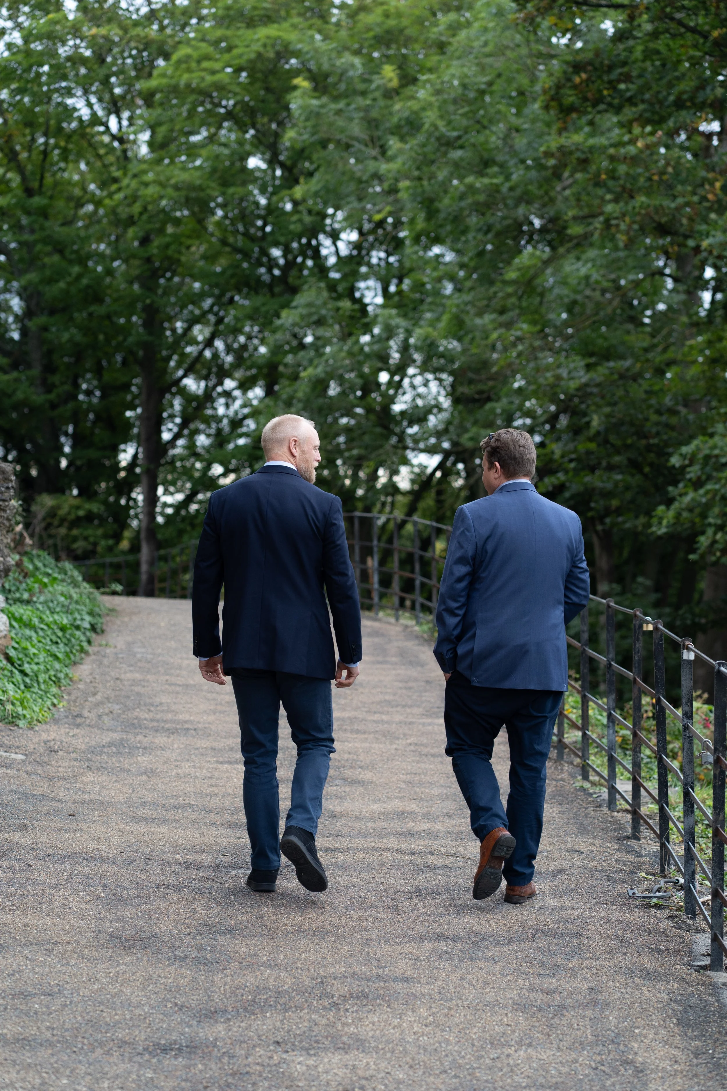 Two men in suits walking along a gravel path in a park with dense green trees.