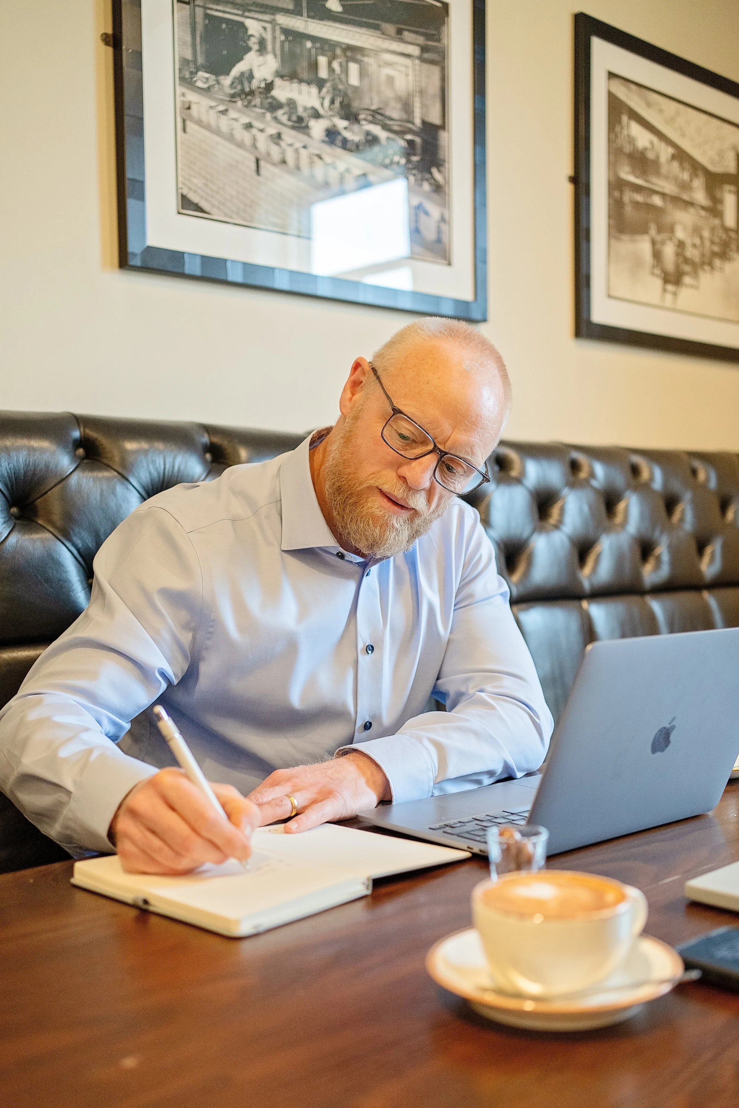 A man with glasses writing in a notebook in front of a laptop in a café or office setting, with framed pictures on the wall behind him and a cup of coffee on the table.
