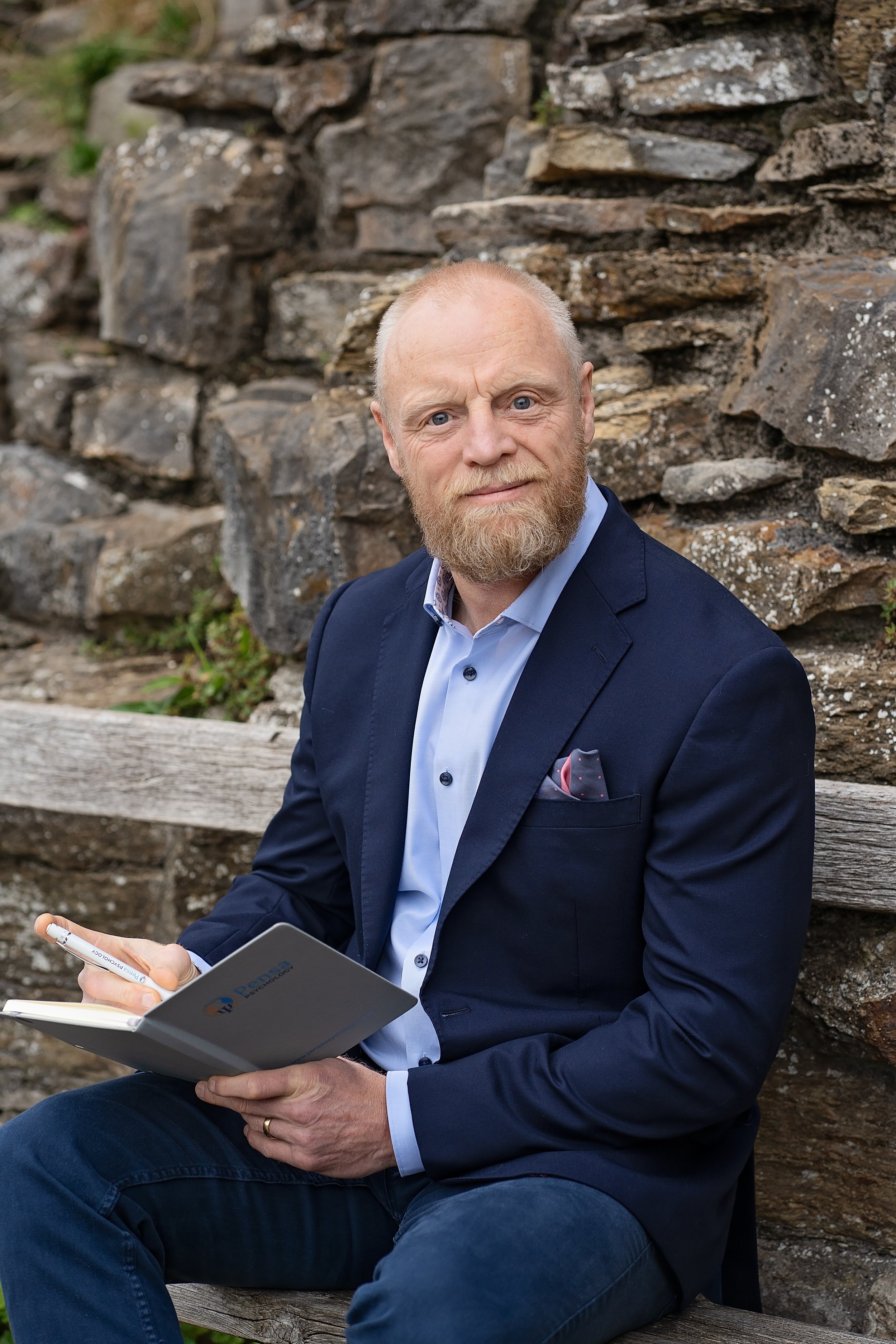 A man with a red beard, wearing a navy blazer and light blue shirt, sitting on a wooden bench in front of a stone wall, holding a notebook and pen, looking at the camera.