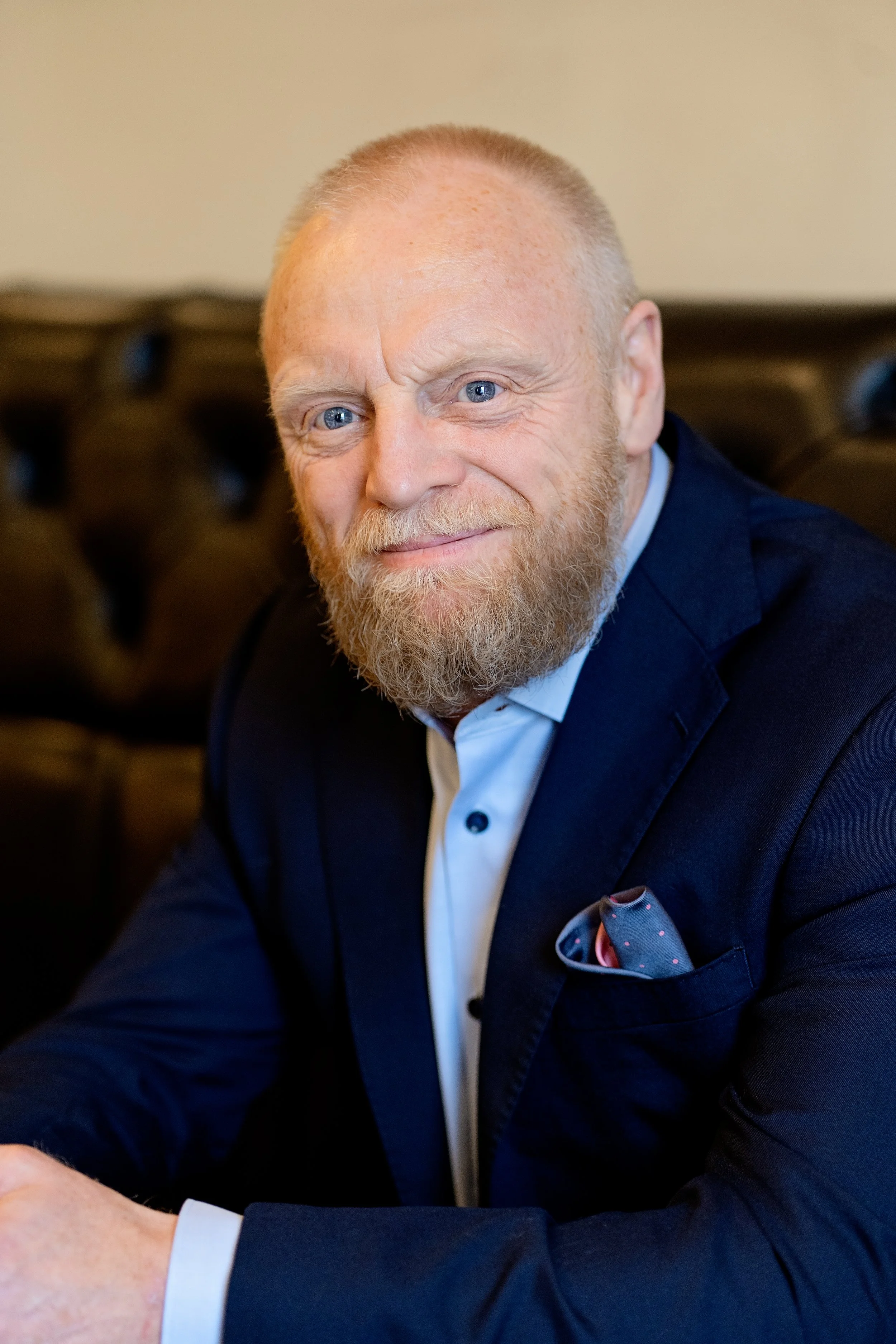 A middle-aged man with a beard and blue eyes, dressed in a navy suit and white shirt, sitting indoors with a dark-colored leather couch in the background.
