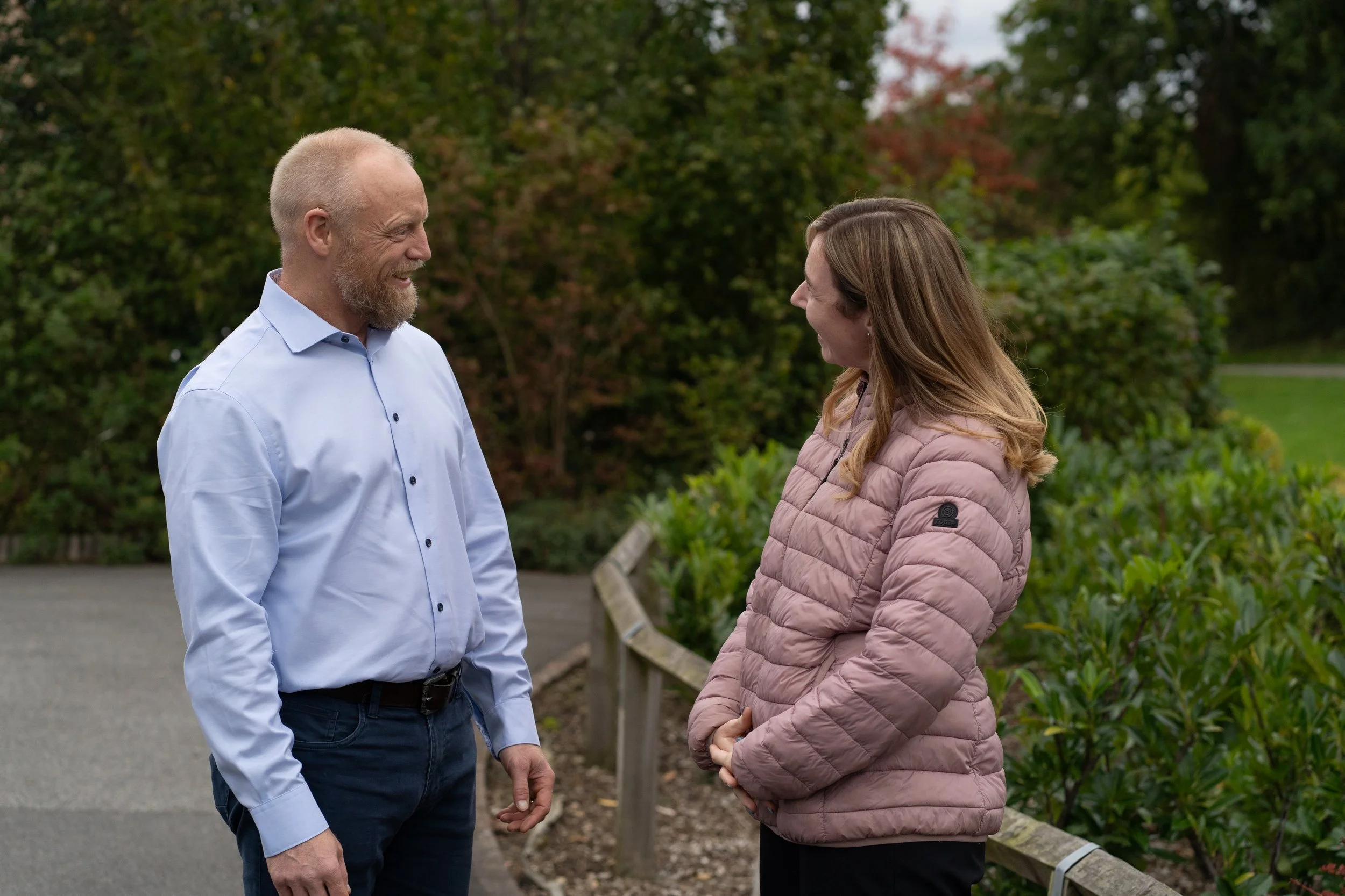 A man and woman are talking outdoors, standing on a pathway with trees and bushes in the background.