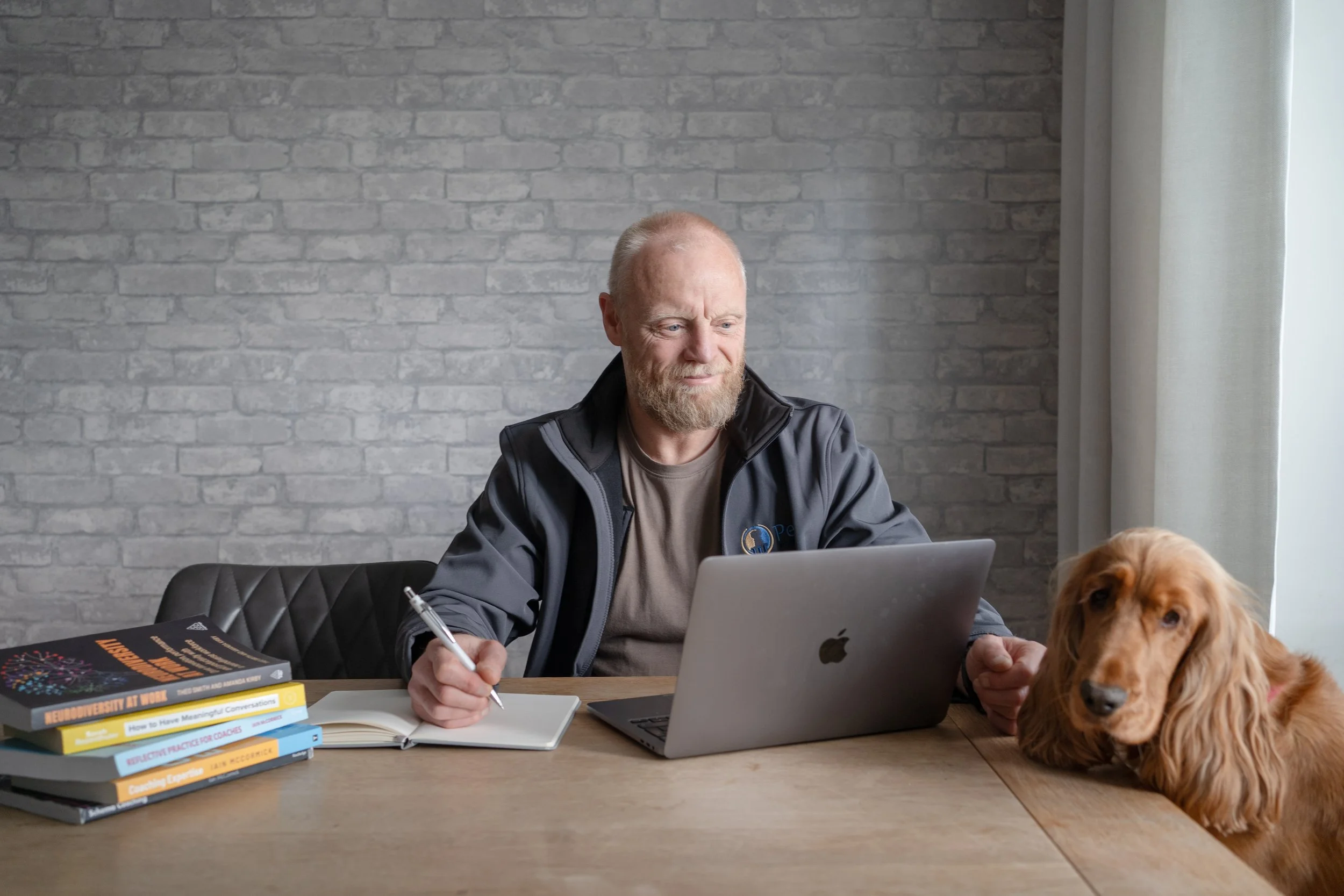 A man working at a desk with a laptop and notebooks, with a dog sitting nearby.