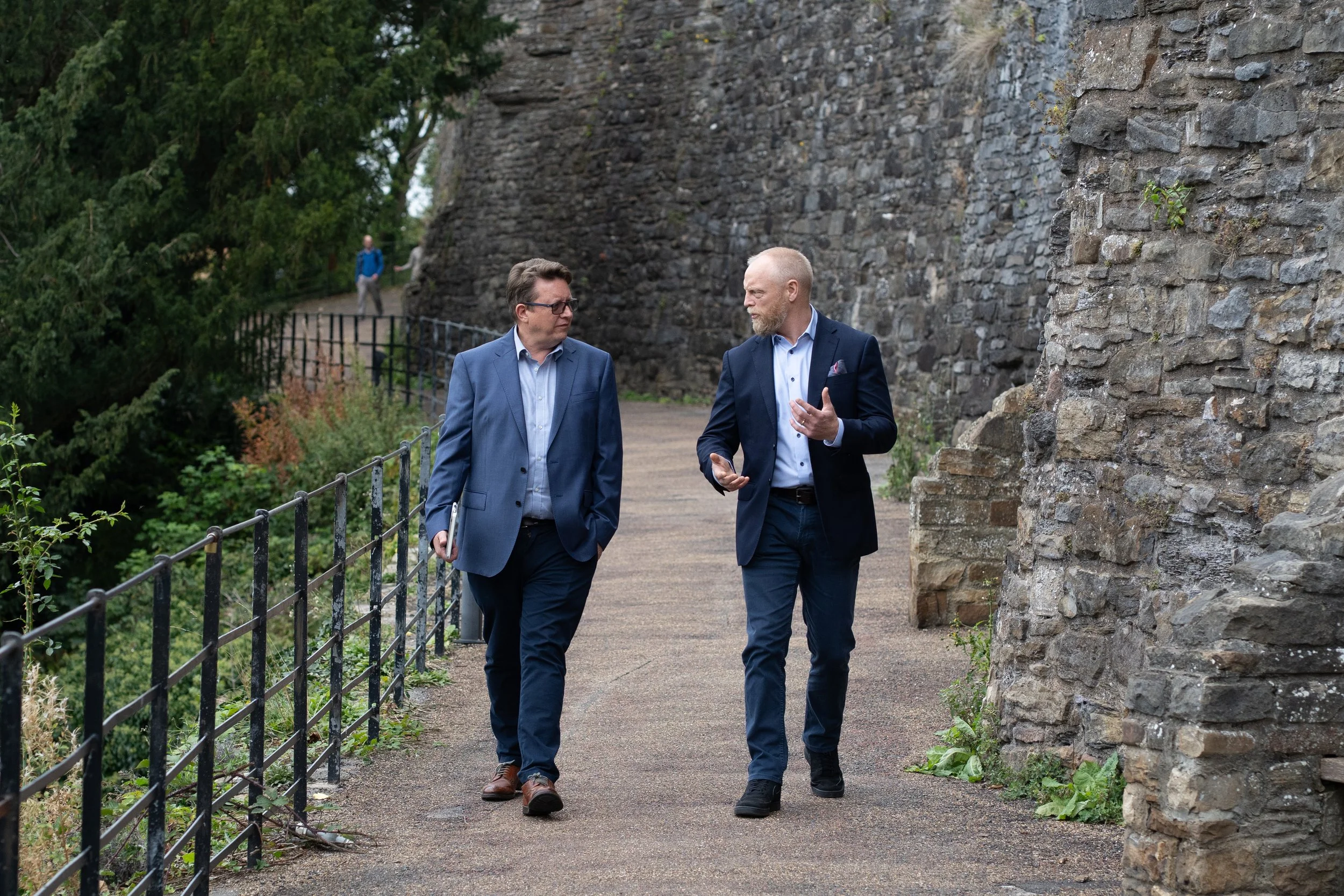 Two men in suits walking and talking along a pathway next to a stone wall, surrounded by greenery.