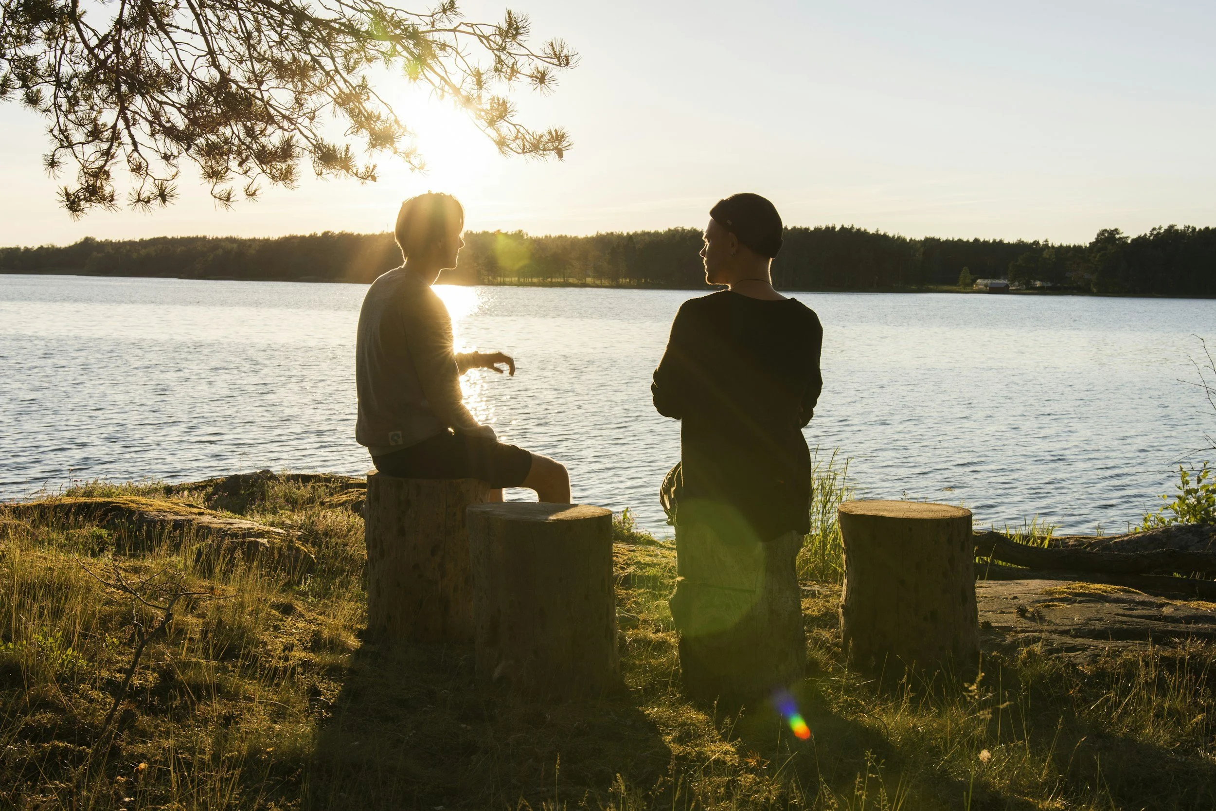 Two people sit on logs by a lake at sunset, talking and enjoying the view, with trees and a building in the background.