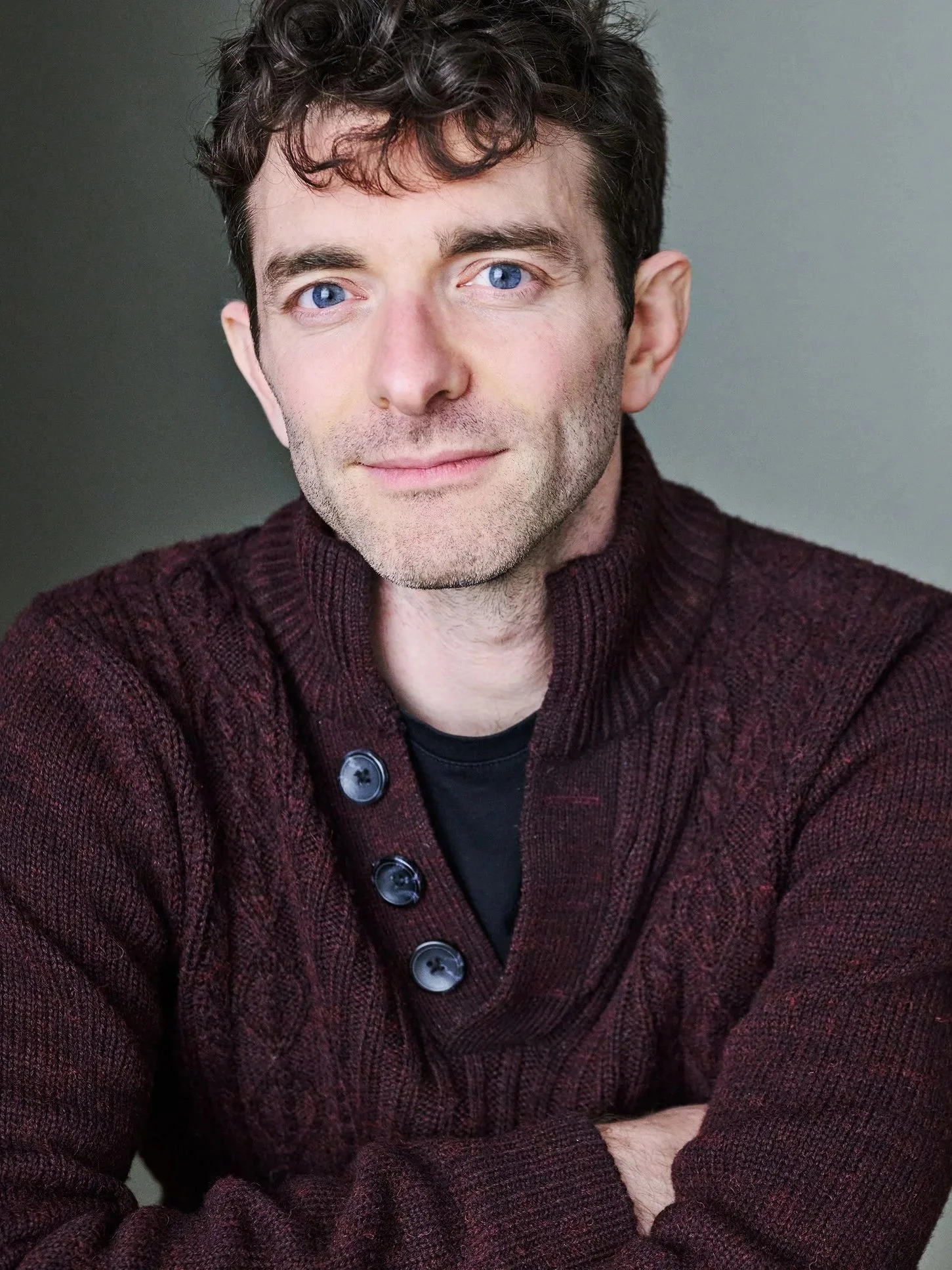 A photo of me. A white man, with curly brown hair and blue eyes wearing a maroon button-up sweater over a black shirt, looking at the camera with a neutral, open expression against a plain background.