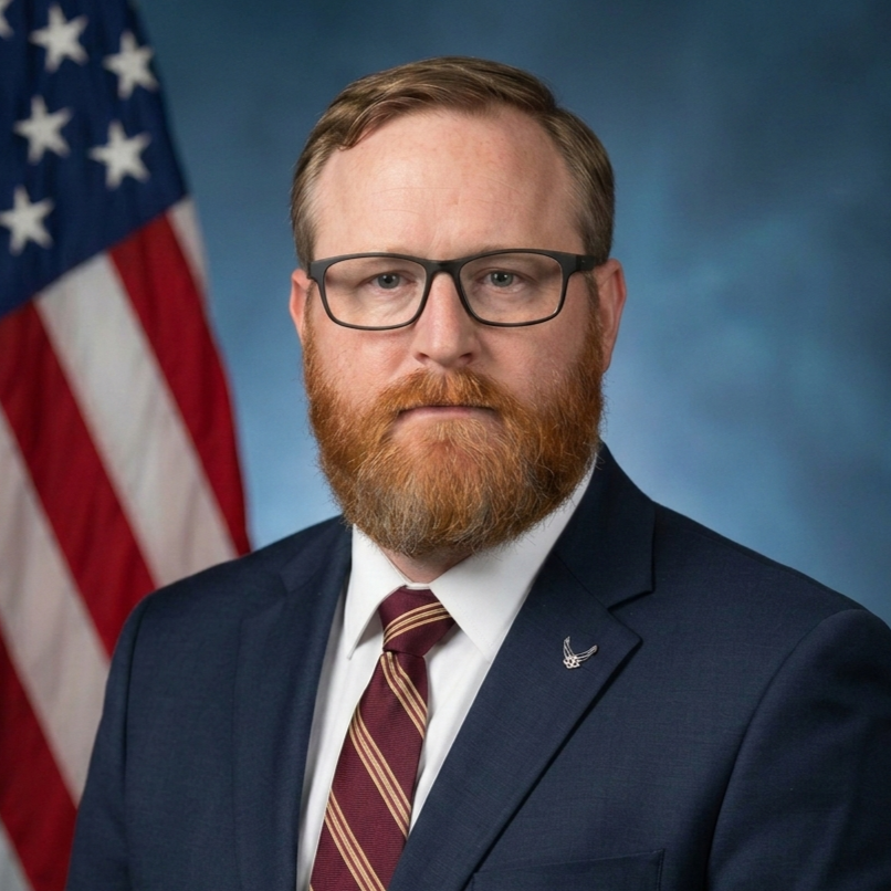 Andrew Naumann. Portrait of a man with short light brown hair, a beard, wearing a dark suit, white shirt, and striped tie, smiling in front of a blue background.