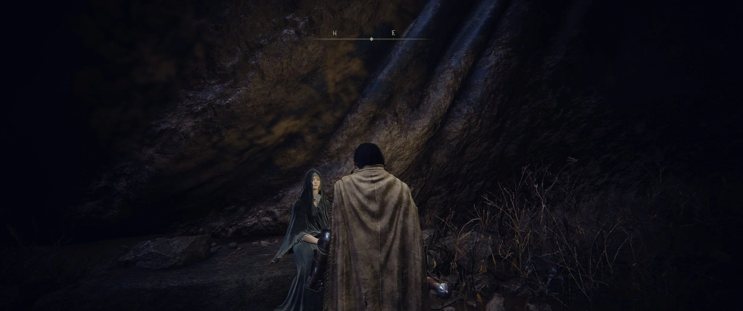 Zwei Menschen in mittelalterlichen Kostümen in einer dunklen Höhle, ein Mann mit Umhang und eine Frau mit dunklem Kleid und Schleier, umgeben von Steinen und trockenen Zweigen.