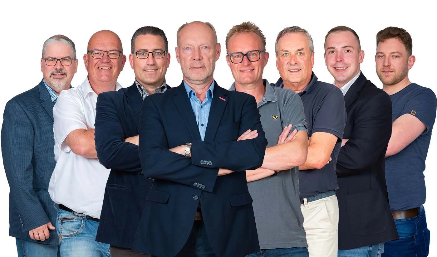 Eine Gruppe von acht Männern in unterschiedlicher Kleidung, einige mit verschränkten Armen, vor einem weißen Hintergrund.