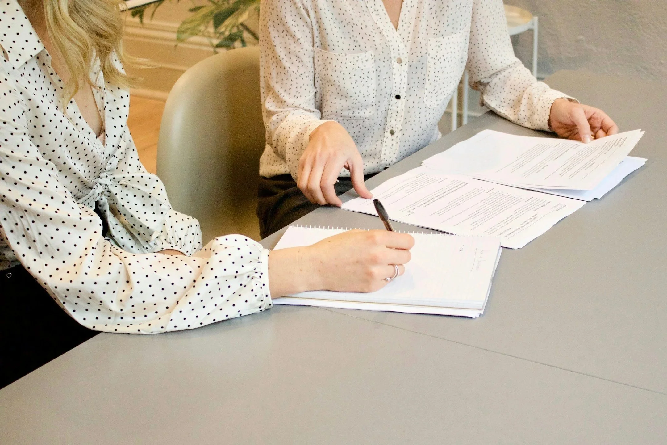Two women sitting at a table with paperwork. One is writing, and the other is pointing at the documents. Both are wearing white blouses with black polka dots.