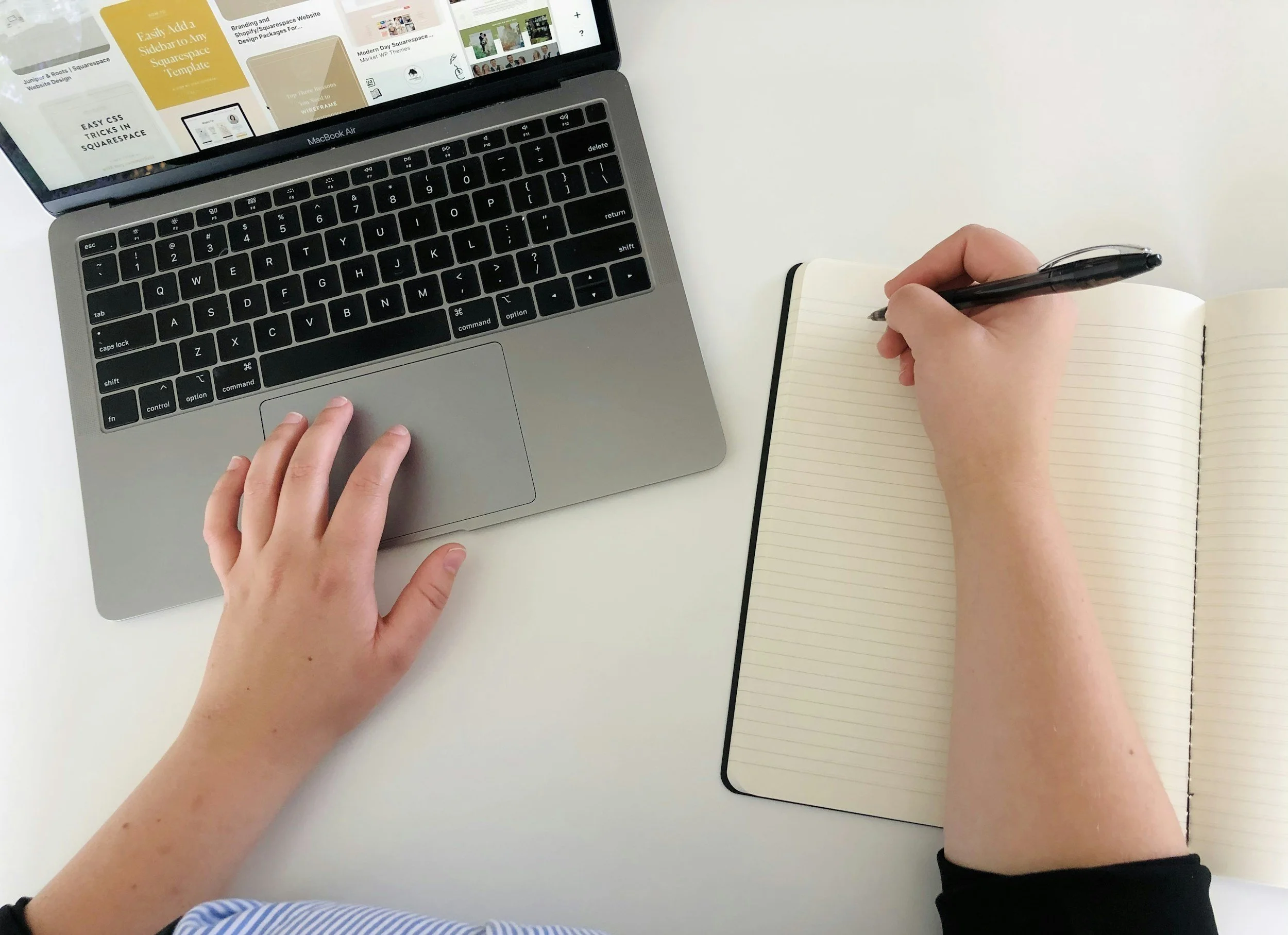 A person sitting at a white table using a MacBook Air laptop with their left hand on the keyboard and small hands holding a black pen and writing in a small, open, lined notebook. There is another closed black notebook on the table nearby.