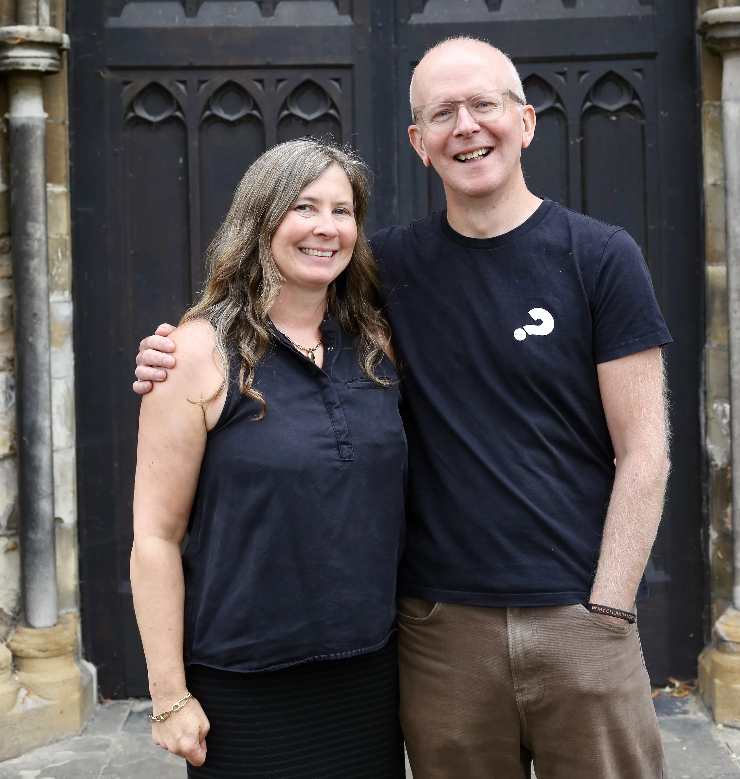 A woman and a man standing close together in front of a wooden door with metal accents, smiling at the camera.