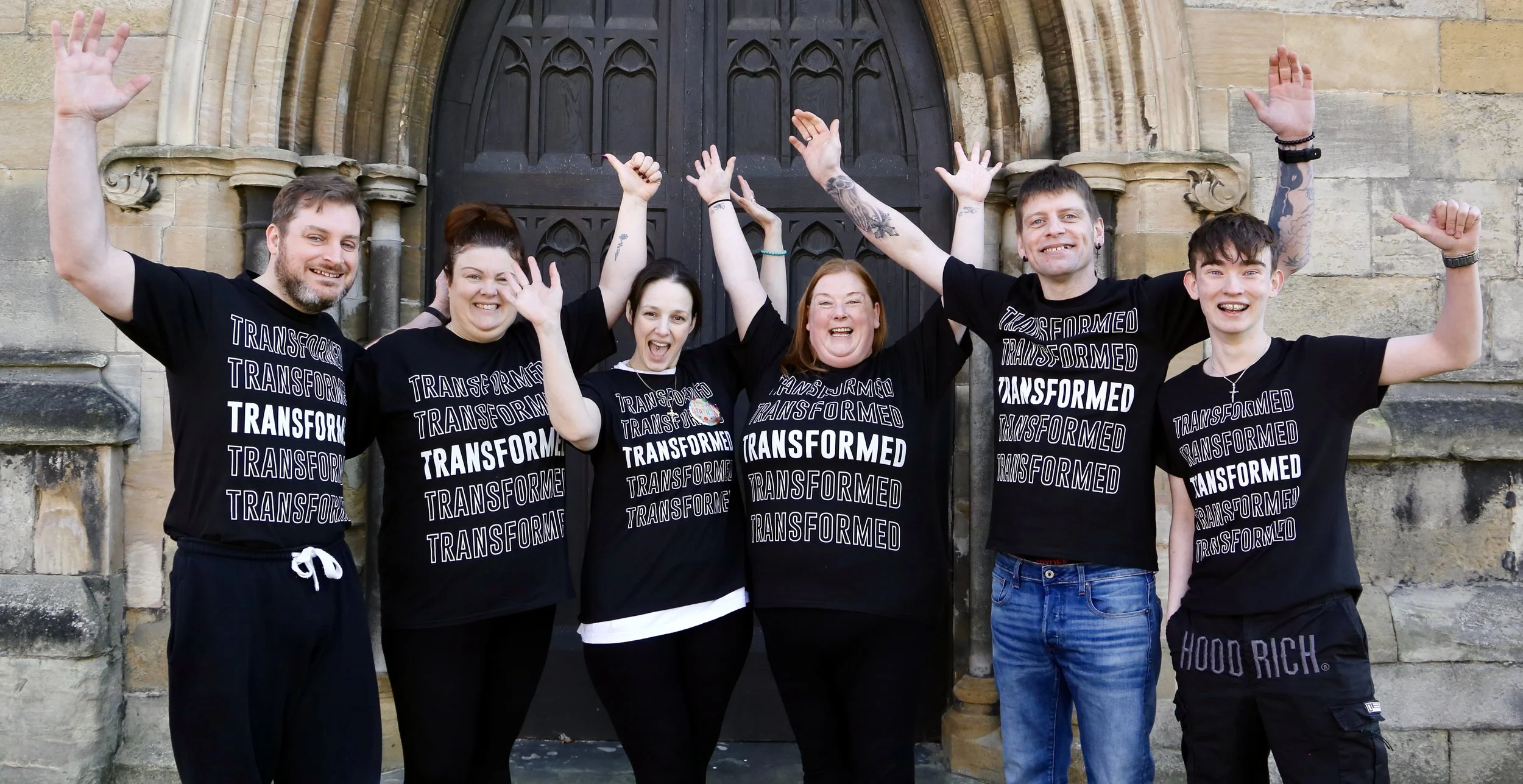 A group of seven people who are being baptised. They are all wearing  T-shirts with the word "TRANSFORMED" printed on,  they are smiling, raising their arms, and appearing joyful and celebratory.