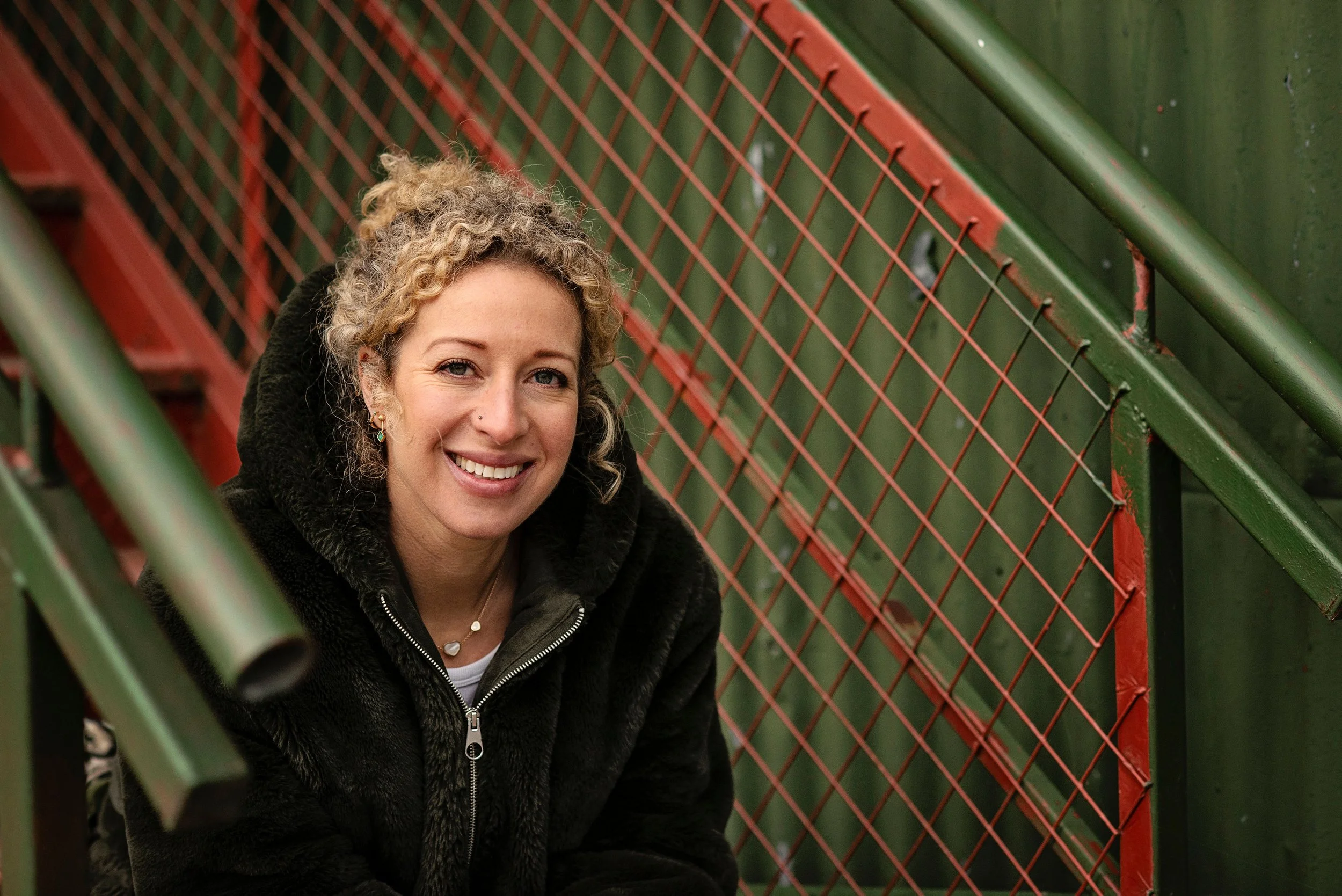Kate Moryoussef smiling warmly, leaning against a green metal staircase railing outdoors.