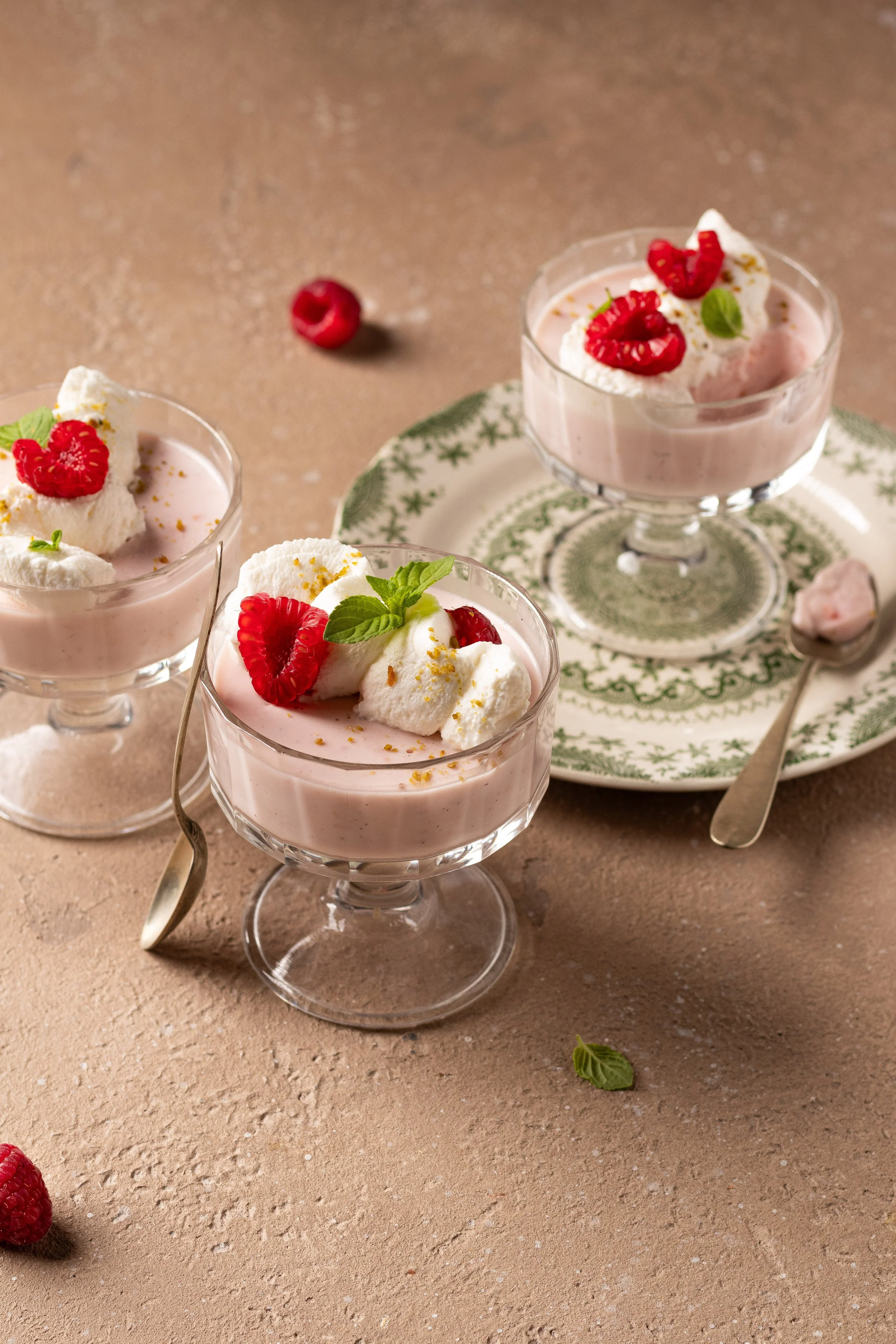 Dessert cremoso rosa con lamponi freschi, meringhe e foglie di menta in coppette di vetro su una tavola di colore beige.