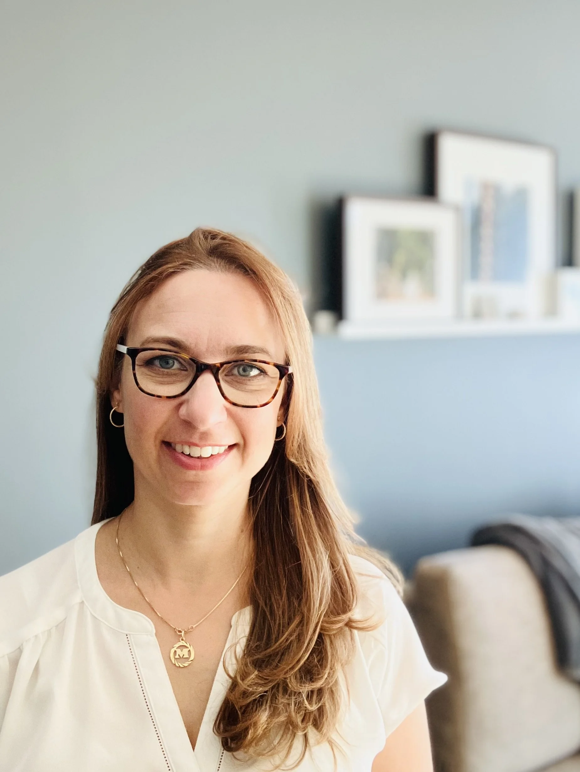 A woman with long light brown hair, eyeglasses, and a white top smiling at the camera in a room with framed pictures on a shelf in the background.