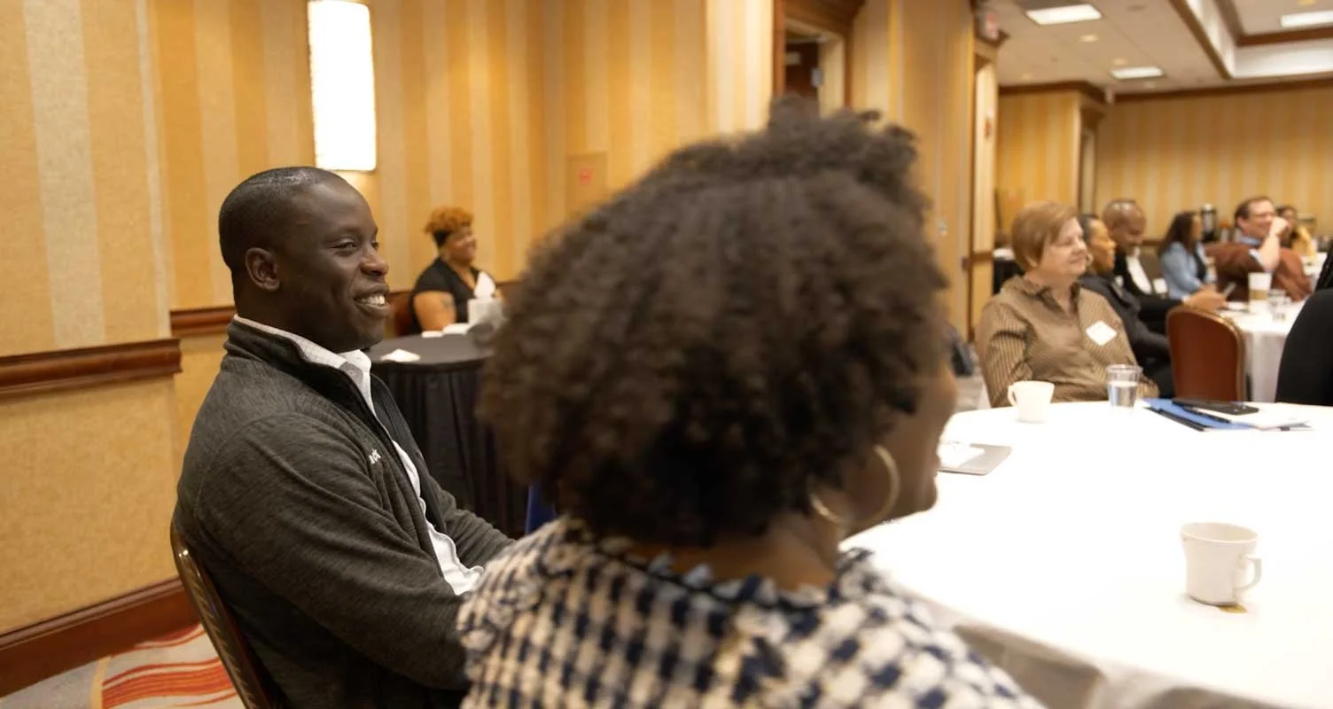 Man in conversation smiling and listening at an event. A woman sits against wall in background.